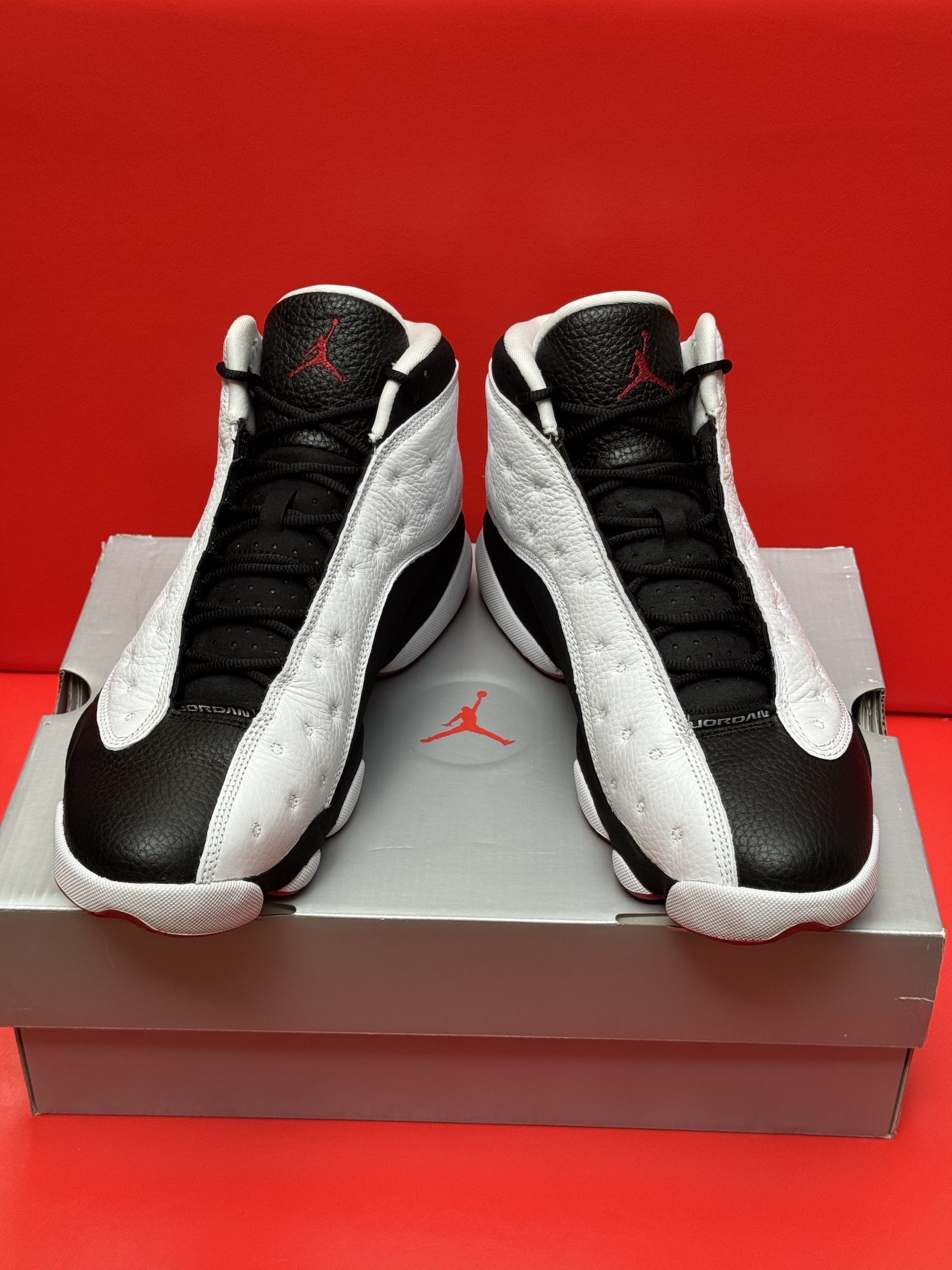 Pair of white, black, and red Jordan 13 sneakers on a grey shoe box, set against a red background.