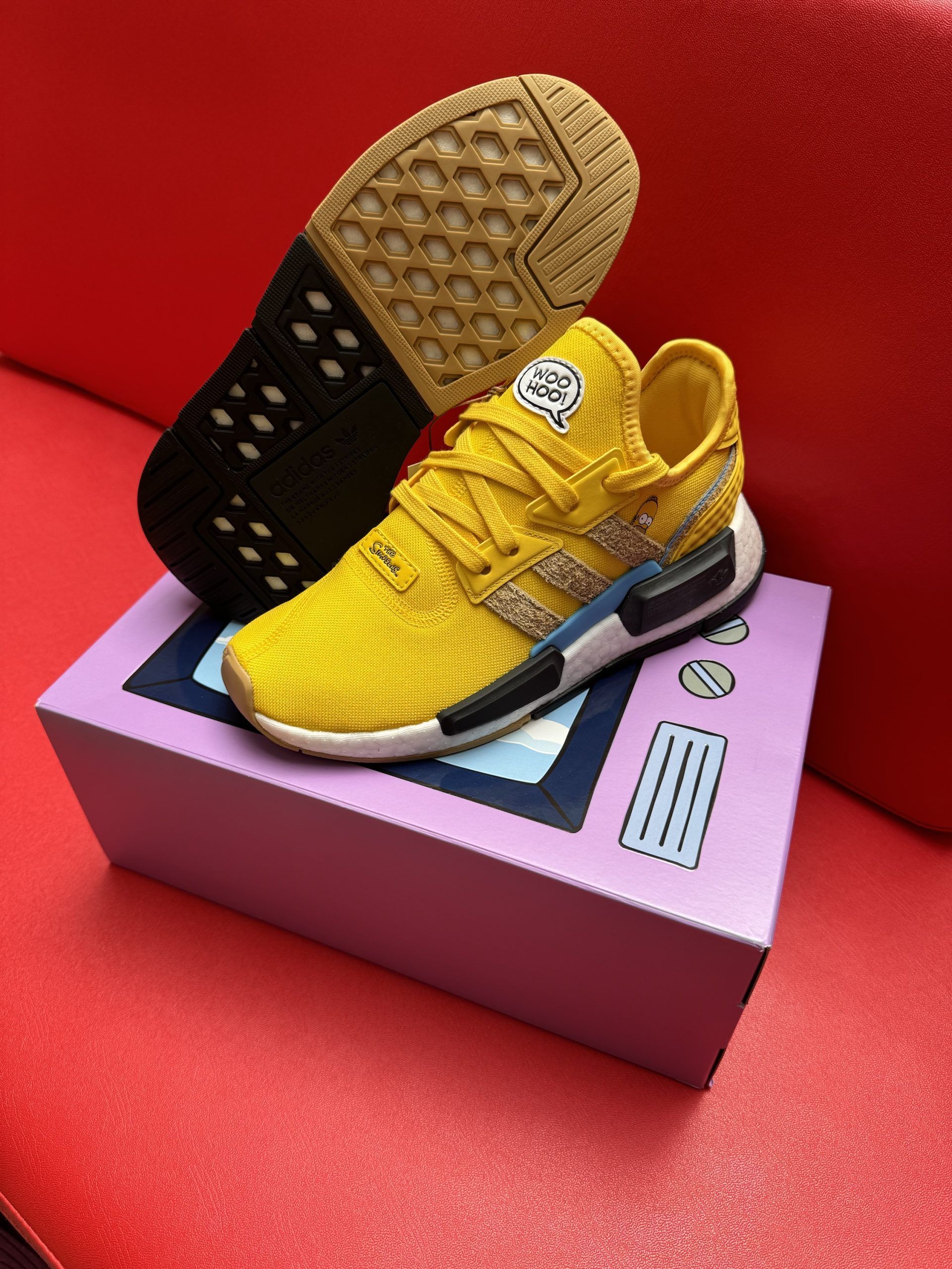 Yellow Adidas sneakers on a purple box with a TV design, set on a red surface.