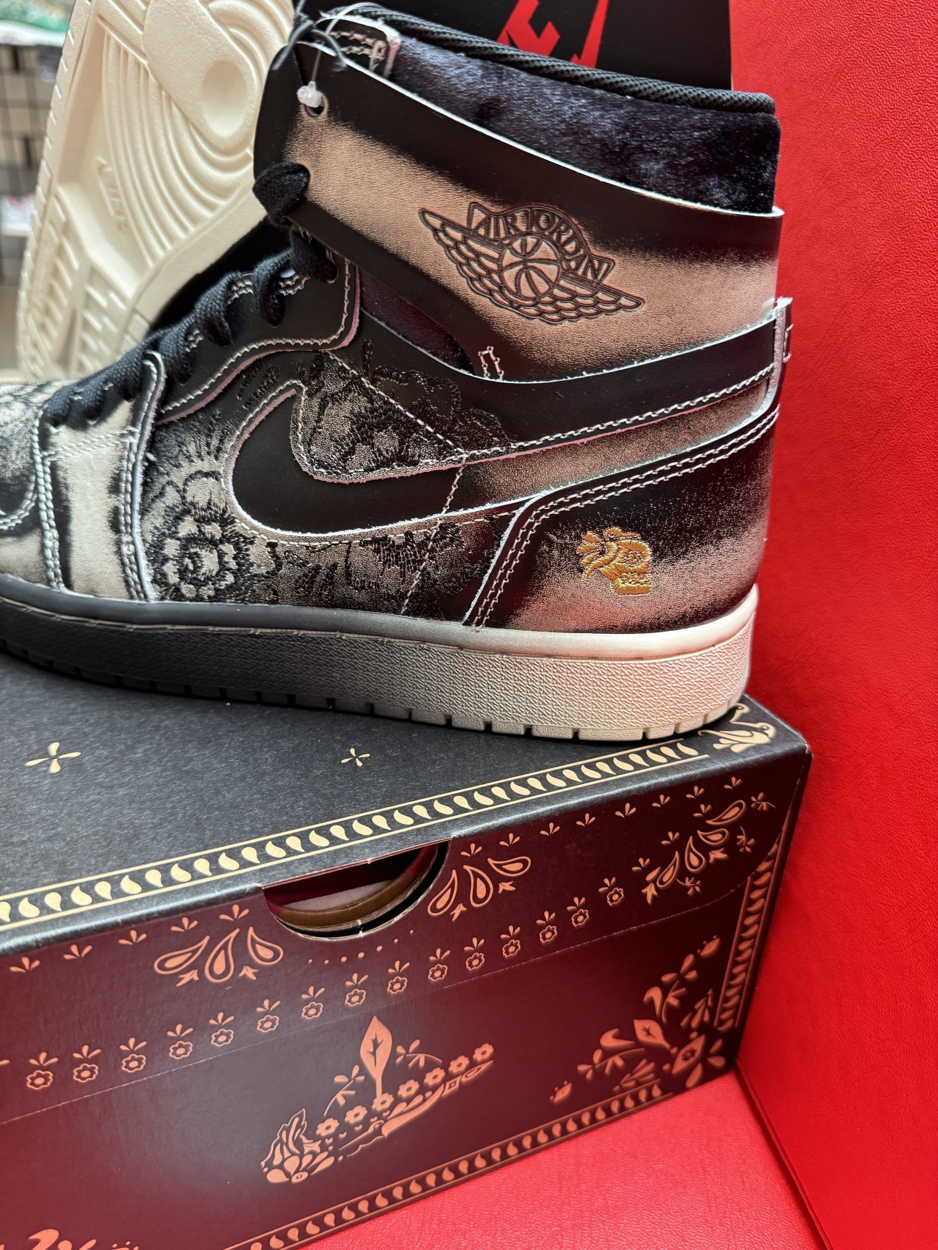 Black and white high-top sneaker with intricate floral pattern on a patterned box. Tan sole.