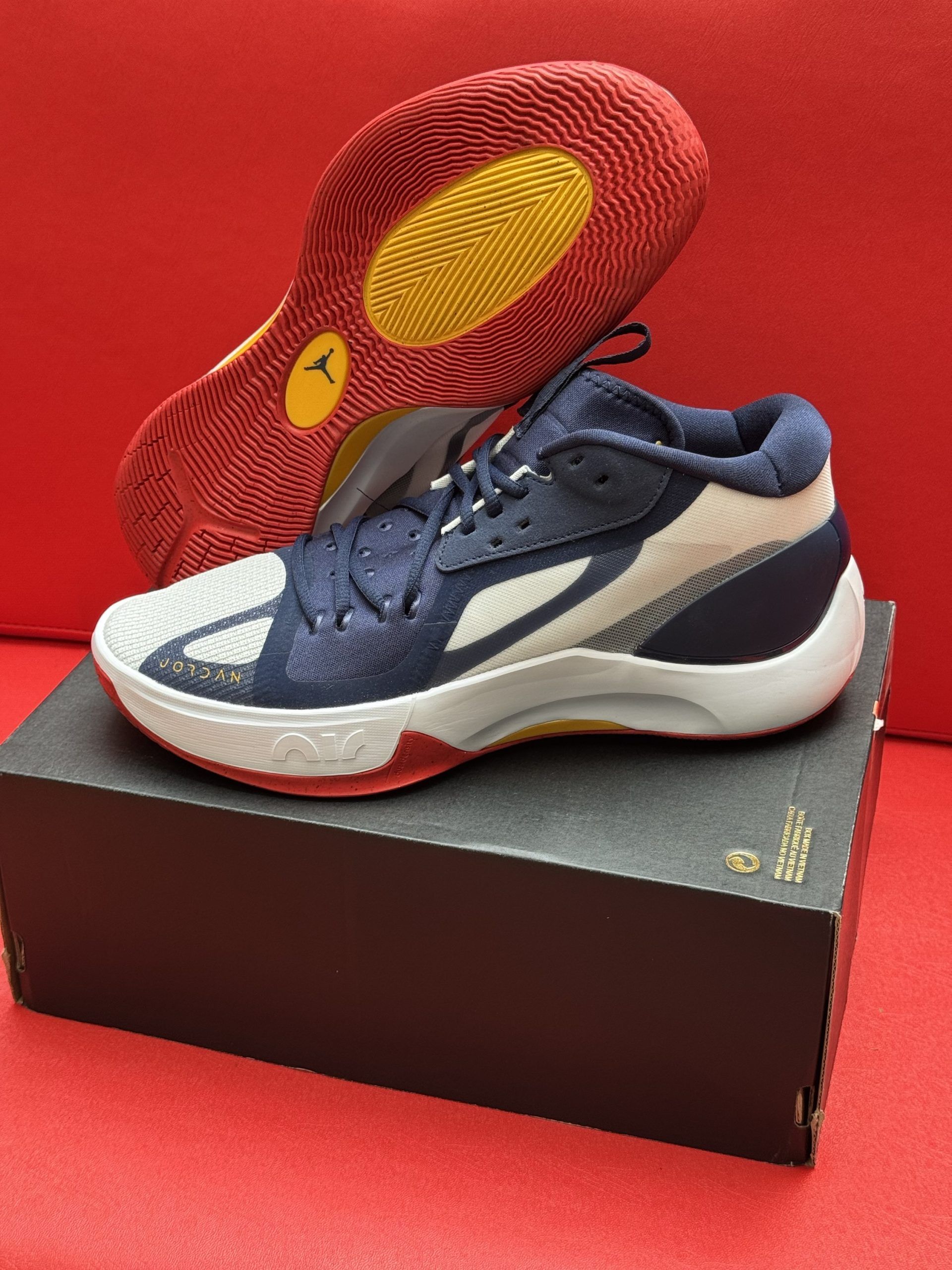 Blue, white, and red basketball shoe, atop a black box, with a red and yellow sole, on a red surface.