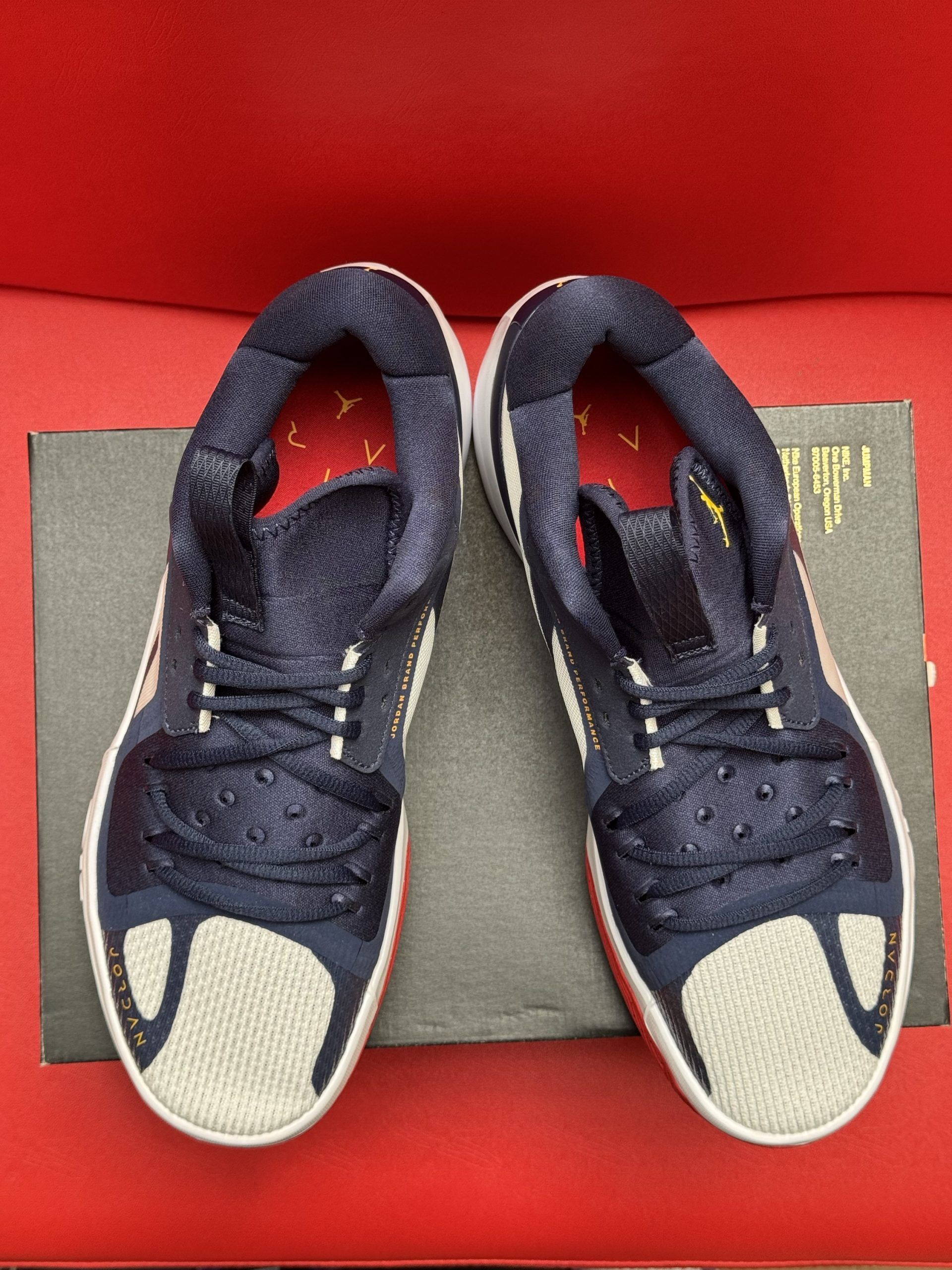 Blue, white, and red basketball shoe, atop a black box, with a red and yellow sole, on a red surface.