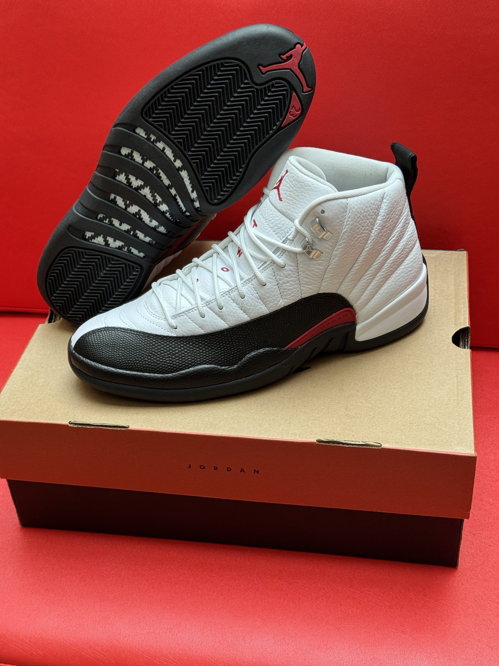 White and black Air Jordan 12 sneakers, red accents, on a cardboard box, red background.