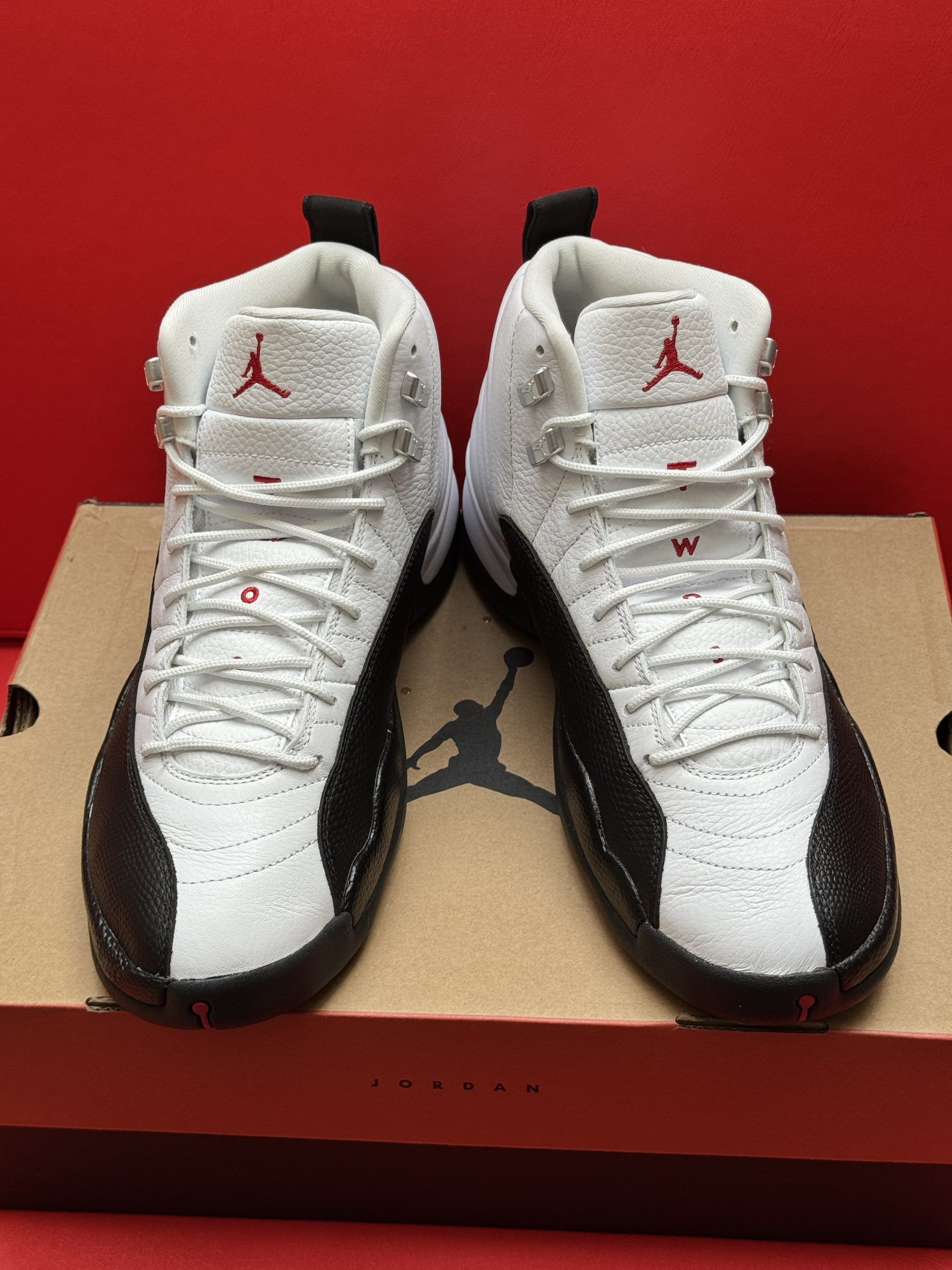 White and black Air Jordan 12 sneakers, red accents, on a cardboard box, red background.
