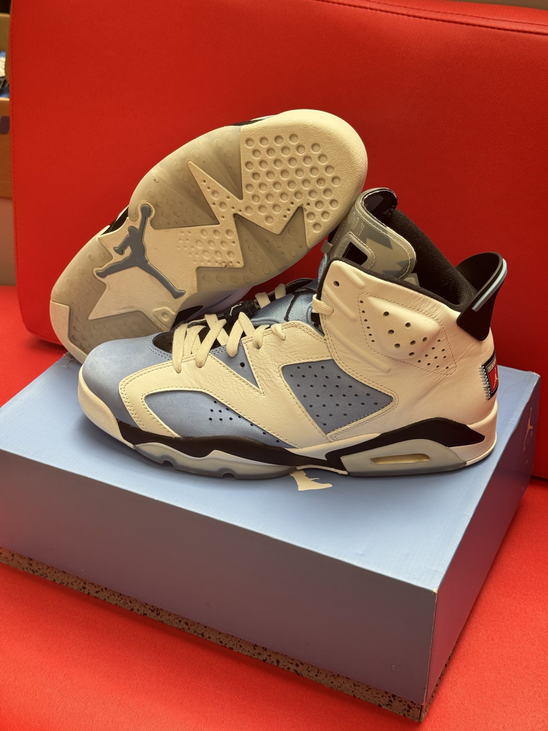 Pair of Air Jordan 6 sneakers in light blue and white, with shoe box on a red surface.