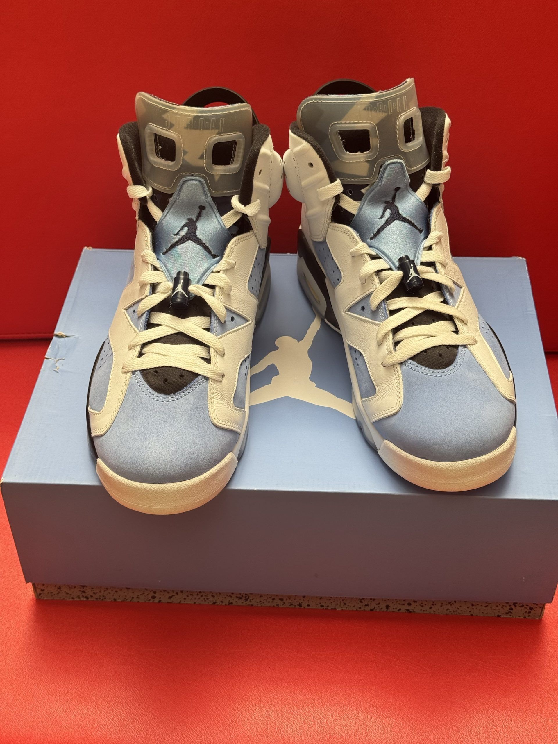 Pair of Air Jordan 6 sneakers in light blue and white, with shoe box on a red surface.