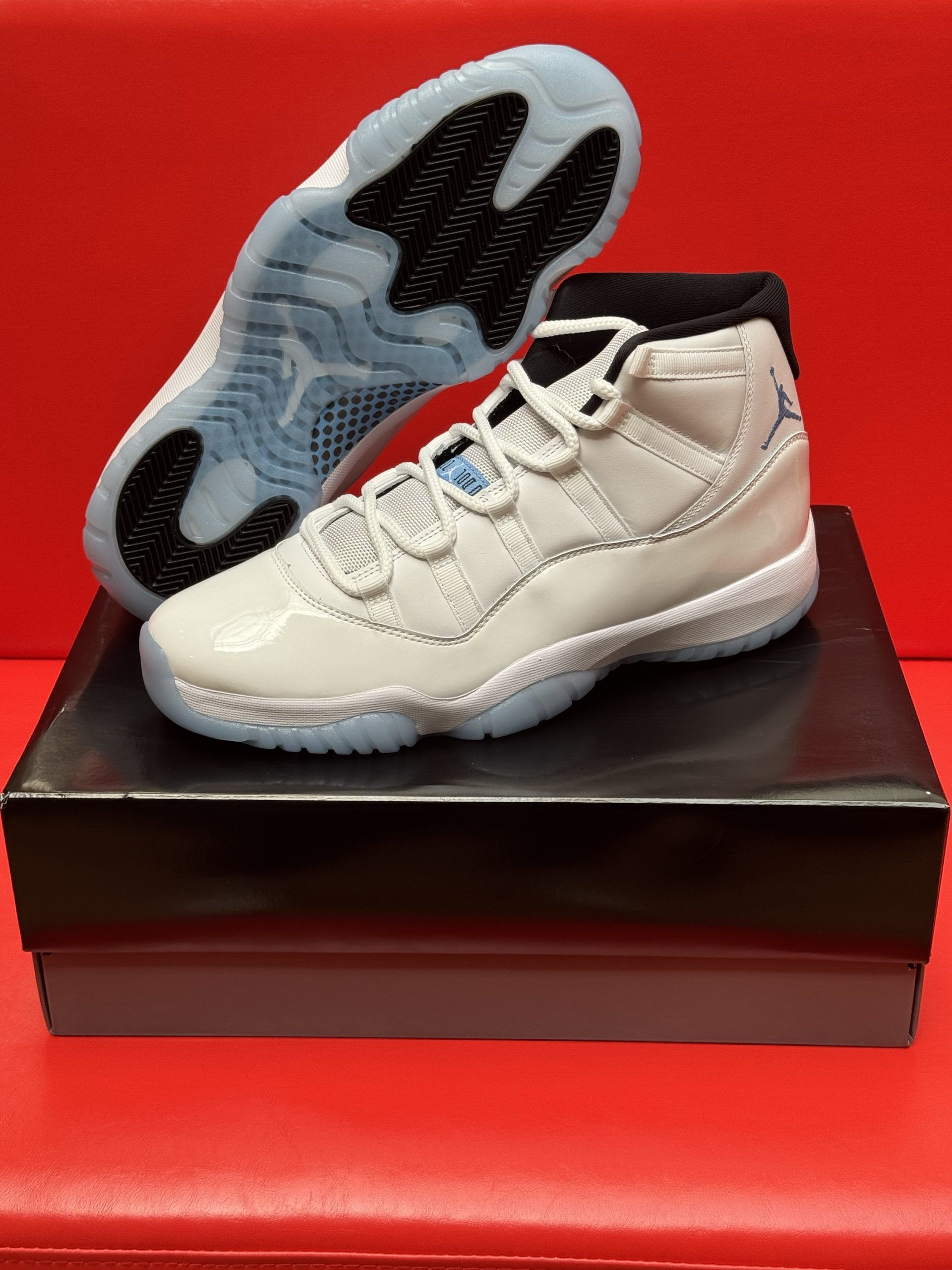 White Air Jordan 11 sneakers on a black box, sole visible, set against a red background.