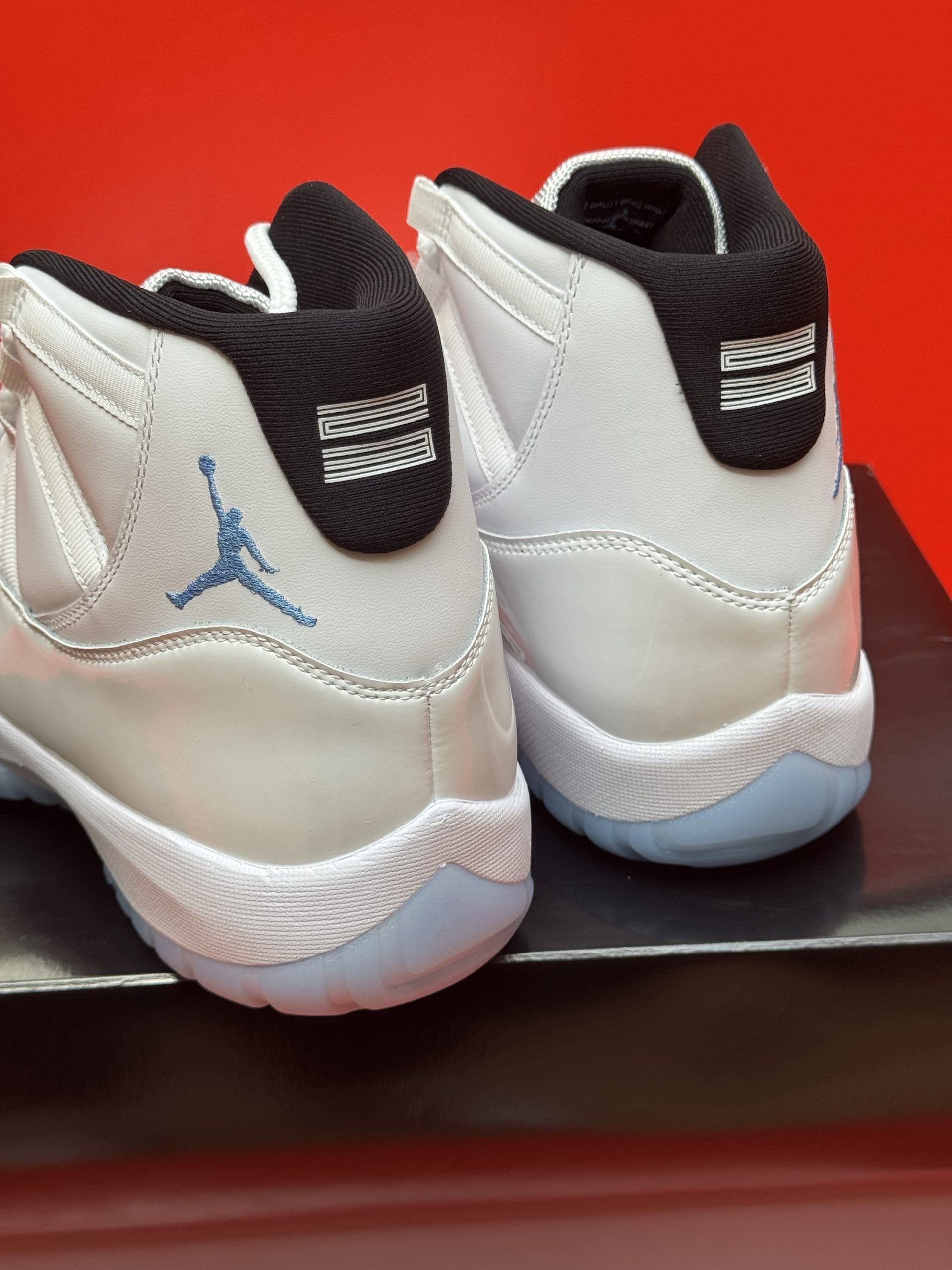 White Air Jordan 11 sneakers on a black box, sole visible, set against a red background.