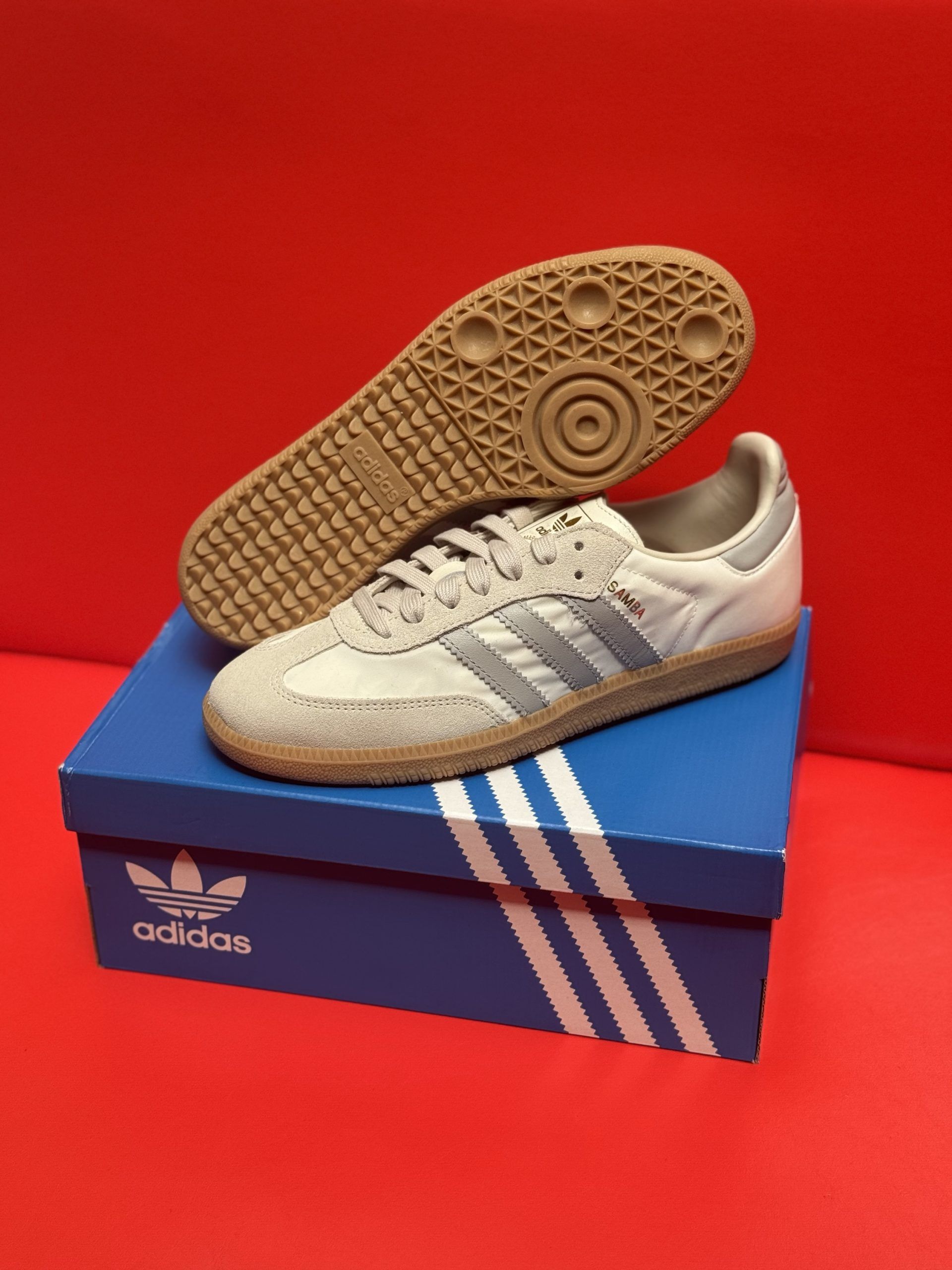 Adidas Samba sneakers, pale gray with silver stripes, on a blue box, sole visible.