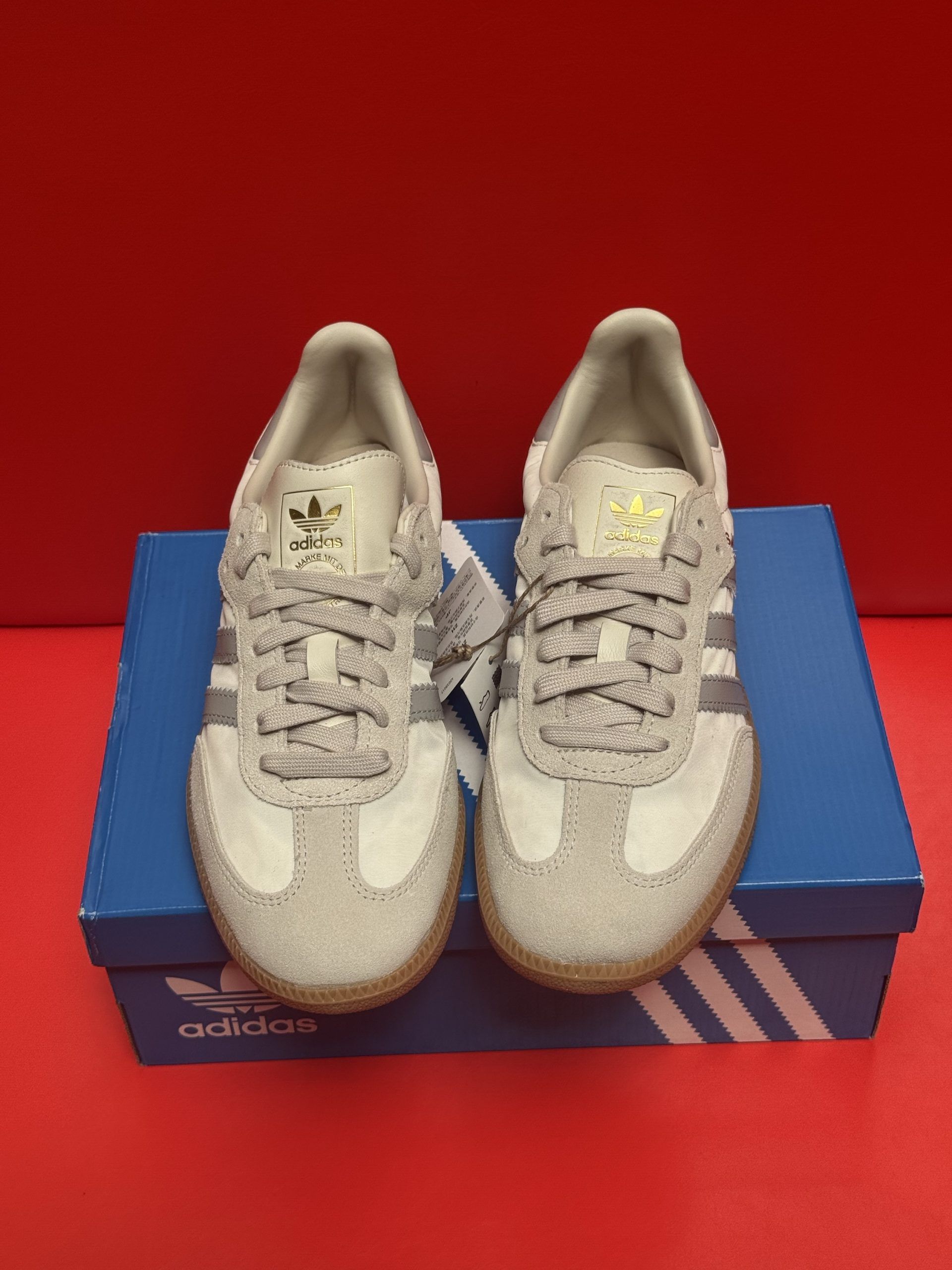 Adidas Samba sneakers, pale gray with silver stripes, on a blue box, sole visible.