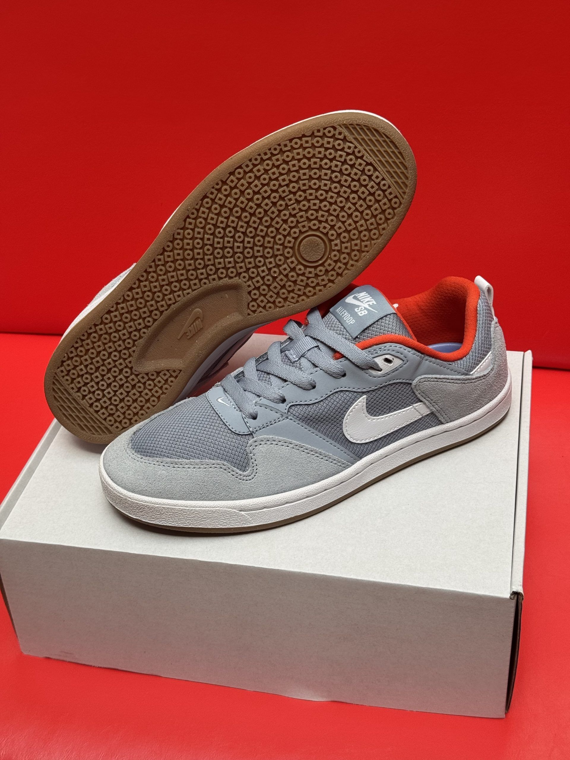 Gray and white Nike sneakers on a white box, one shoe's sole visible, set against a red background.