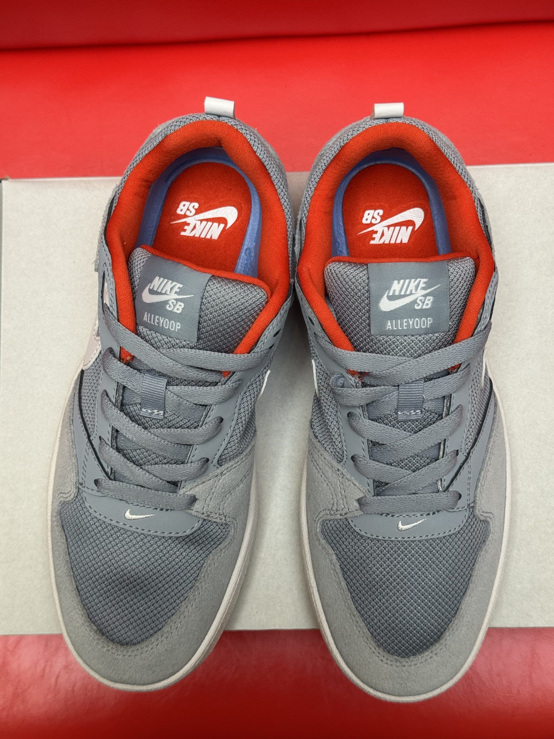 Gray and white Nike sneakers on a white box, one shoe's sole visible, set against a red background.