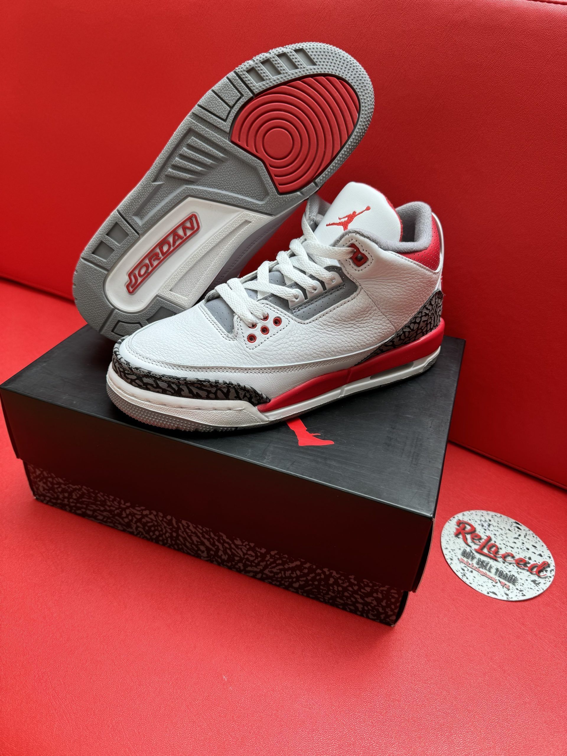 Air Jordan 3 sneakers, white and red, on a black box, with the sole visible. Red background.