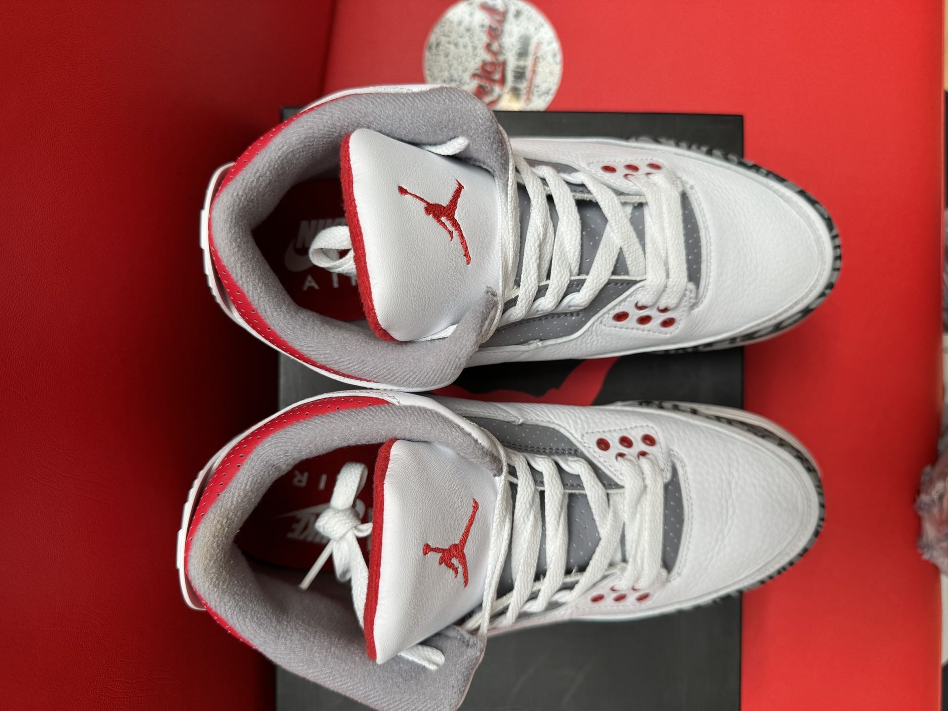 Air Jordan 3 sneakers, white and red, on a black box, with the sole visible. Red background.