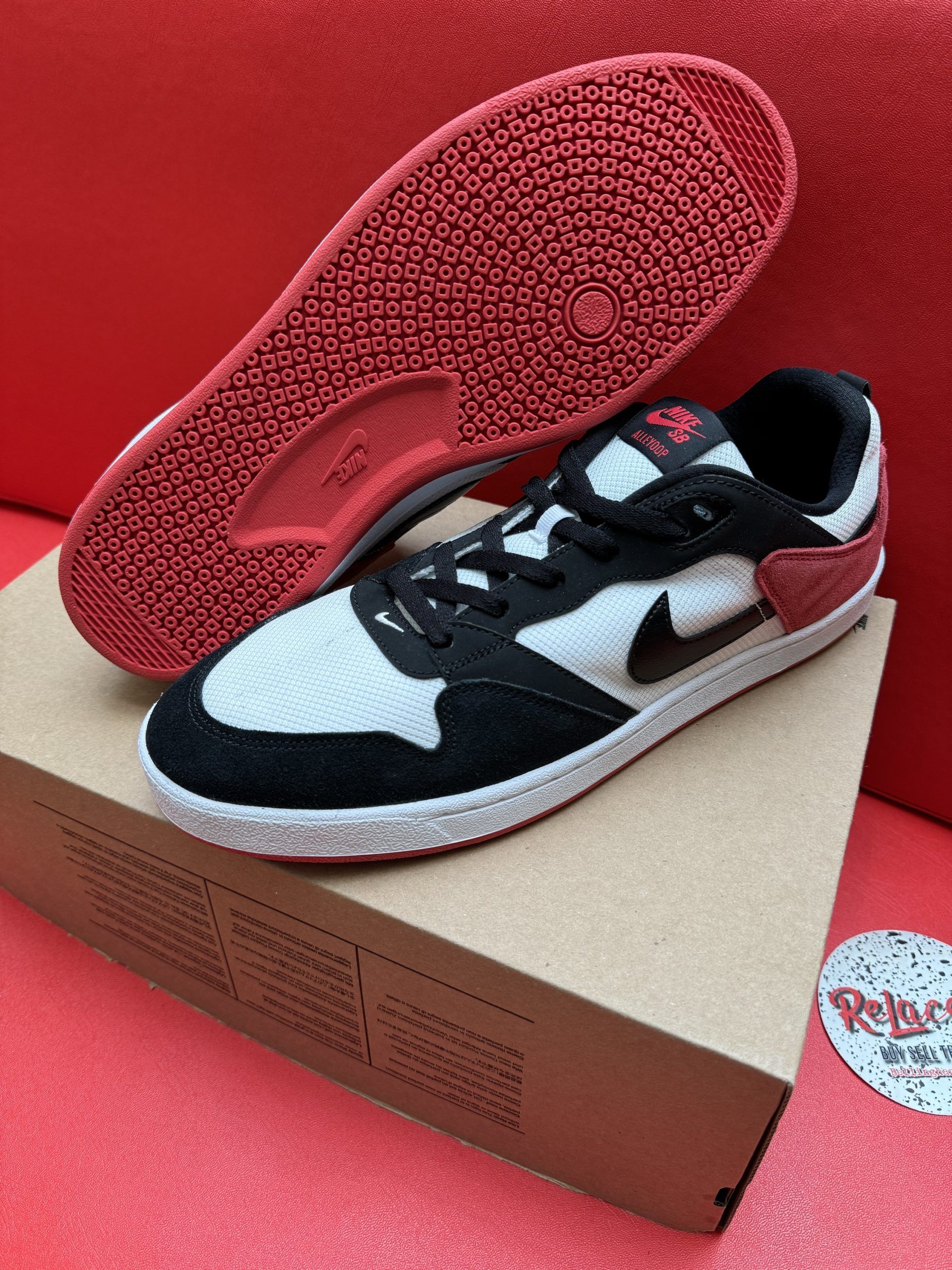 Nike skate shoe, black and white with red accents, on a cardboard box against a red backdrop.