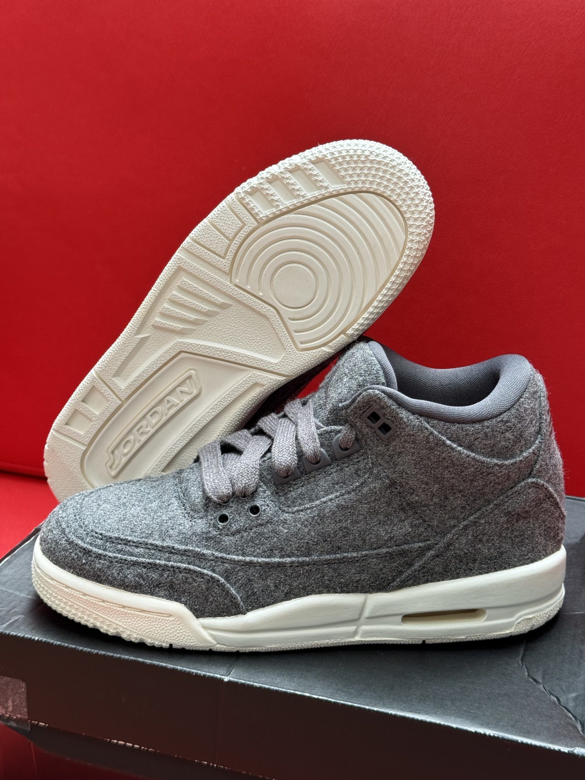 Grey wool Jordan 3 sneakers on a black box, with the sole displayed against a red background.