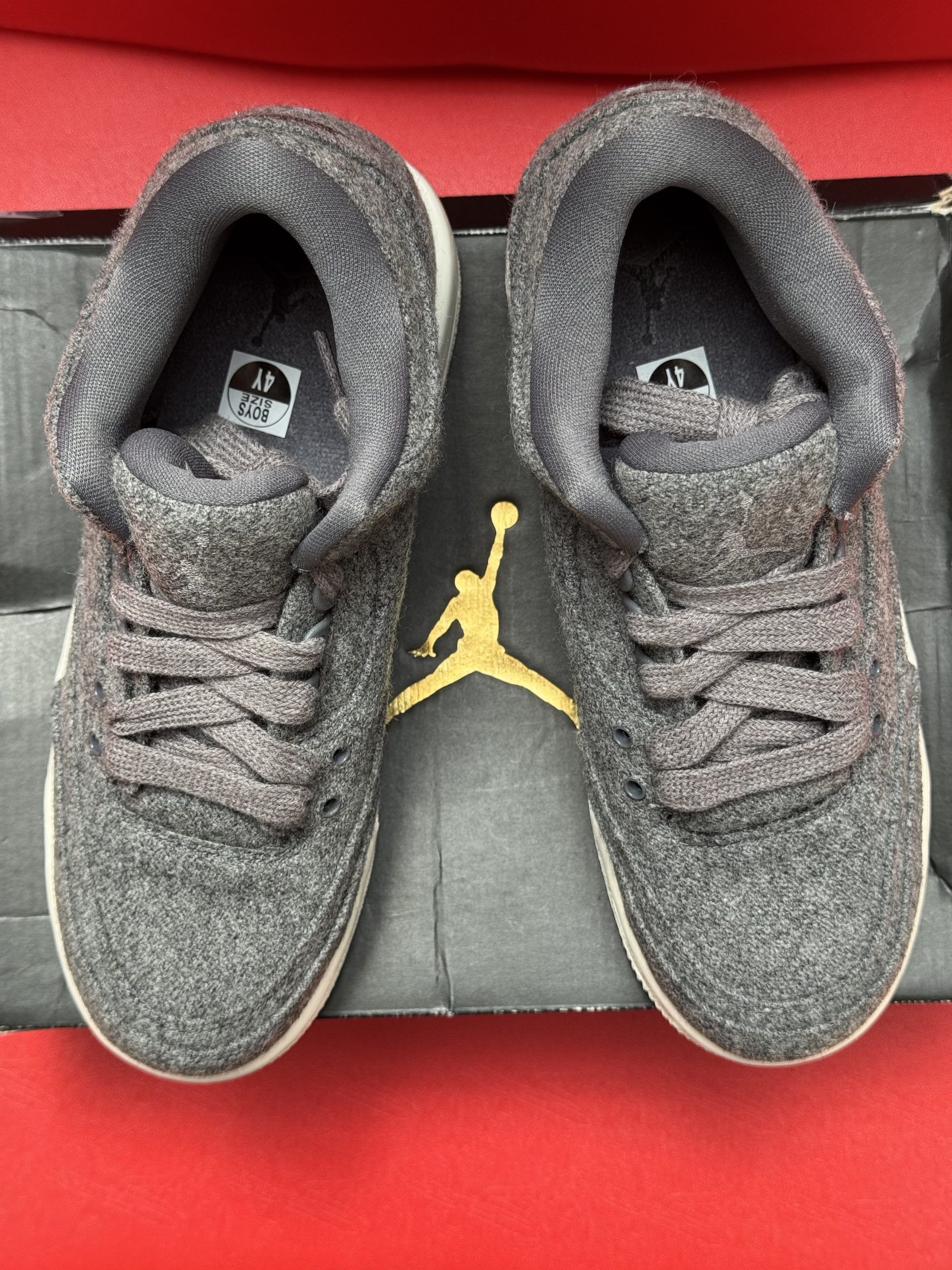 Grey wool Jordan 3 sneakers on a black box, with the sole displayed against a red background.