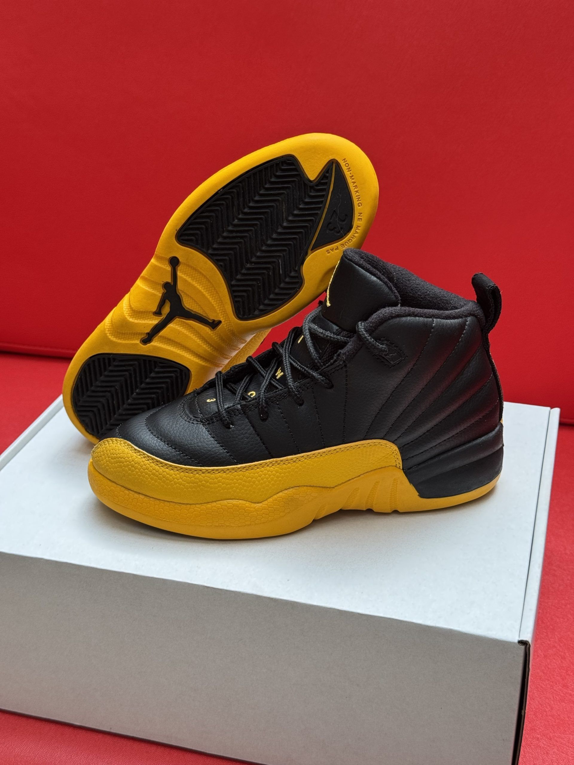Black and yellow Air Jordan 12 sneakers, one on its side revealing yellow sole with black accents, on white box, red background.