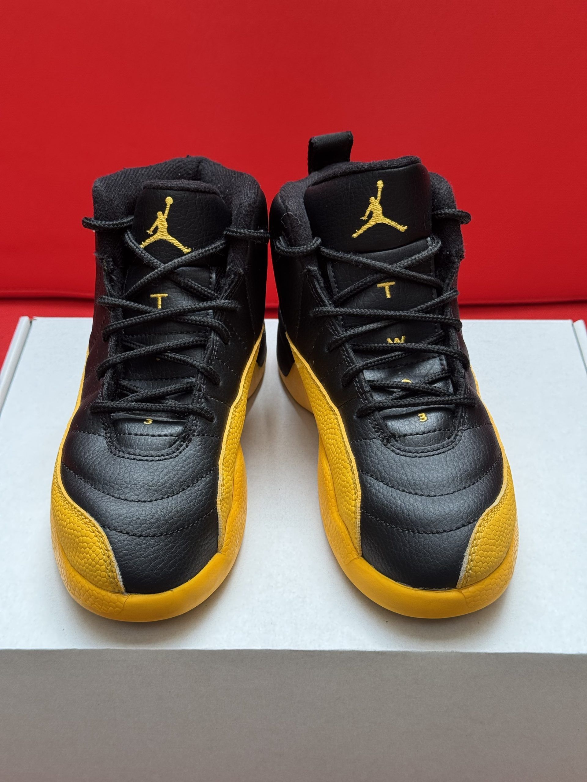 Black and yellow Air Jordan 12 sneakers, one on its side revealing yellow sole with black accents, on white box, red background.