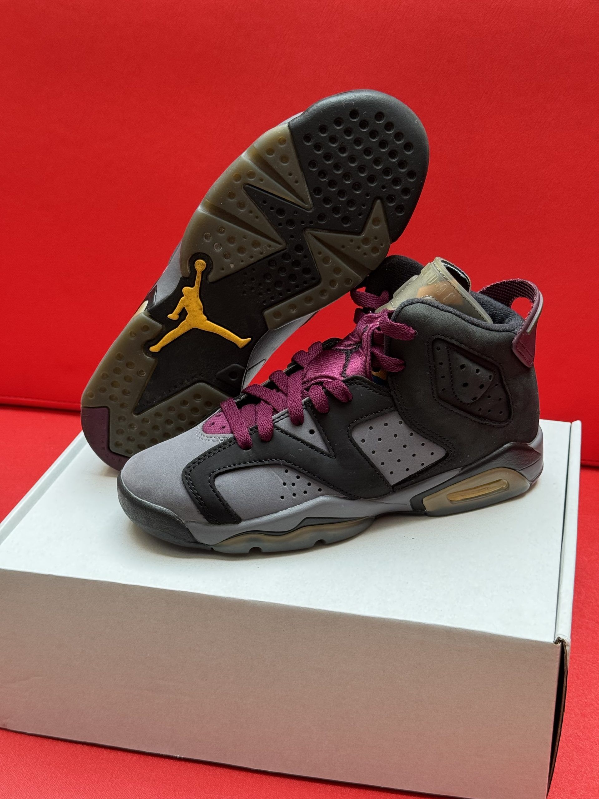 Air Jordan 6 sneakers in gray, black, and maroon, with a brown sole, on a white box against a red backdrop.