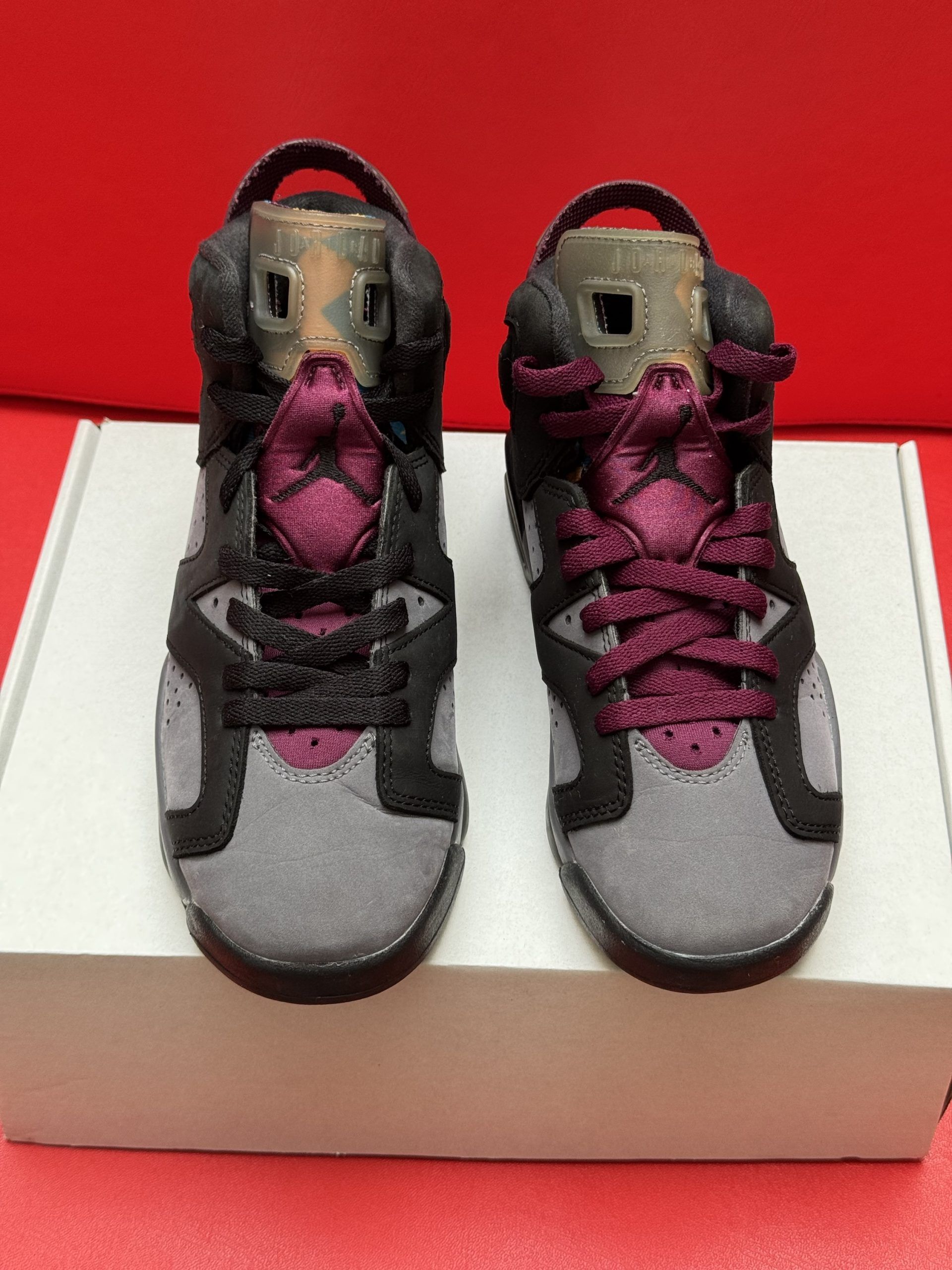Air Jordan 6 sneakers in gray, black, and maroon, with a brown sole, on a white box against a red backdrop.