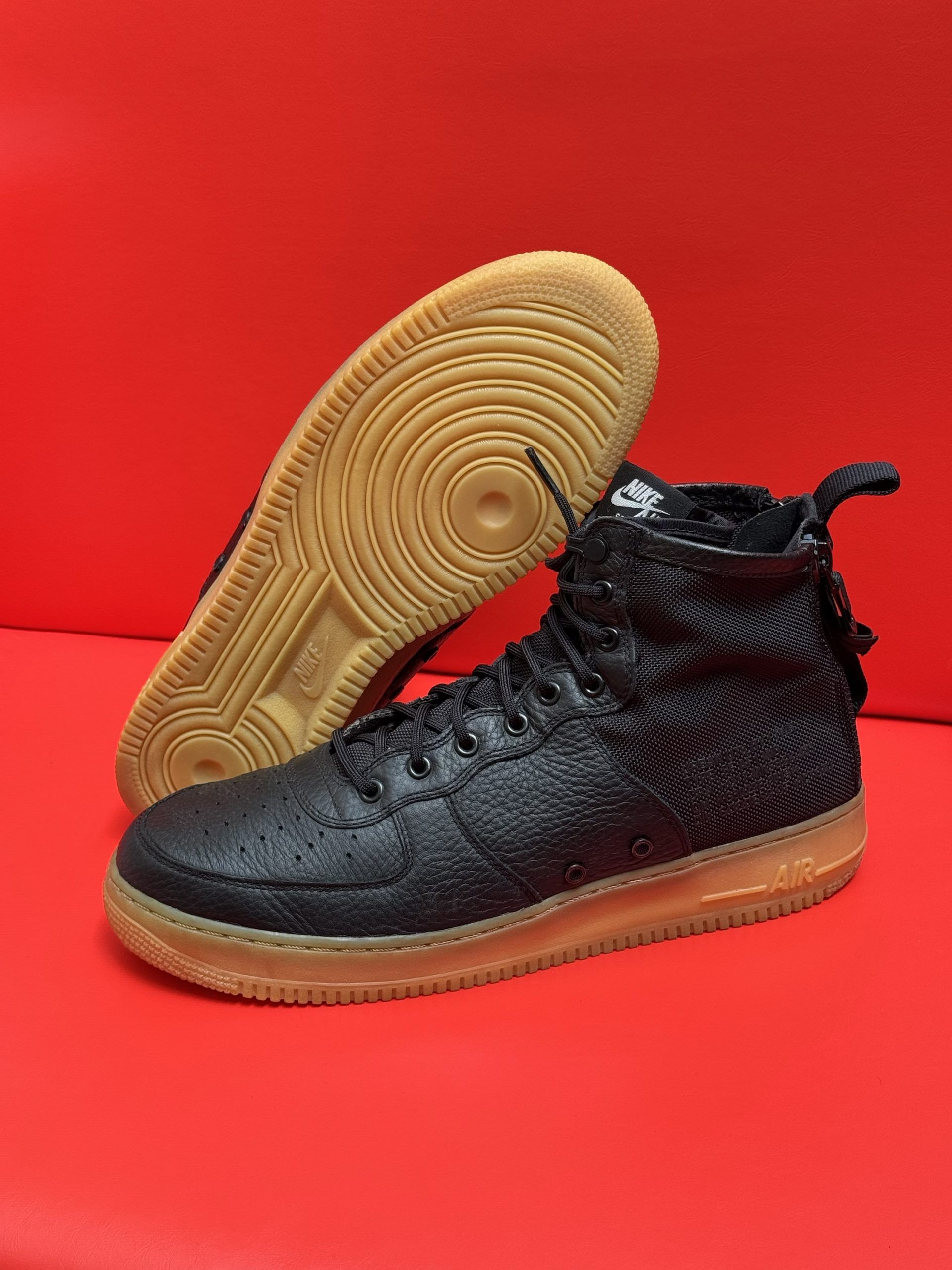 Black high-top sneakers with textured uppers, brown soles, and a red background. One shoe is flipped over to show the sole.