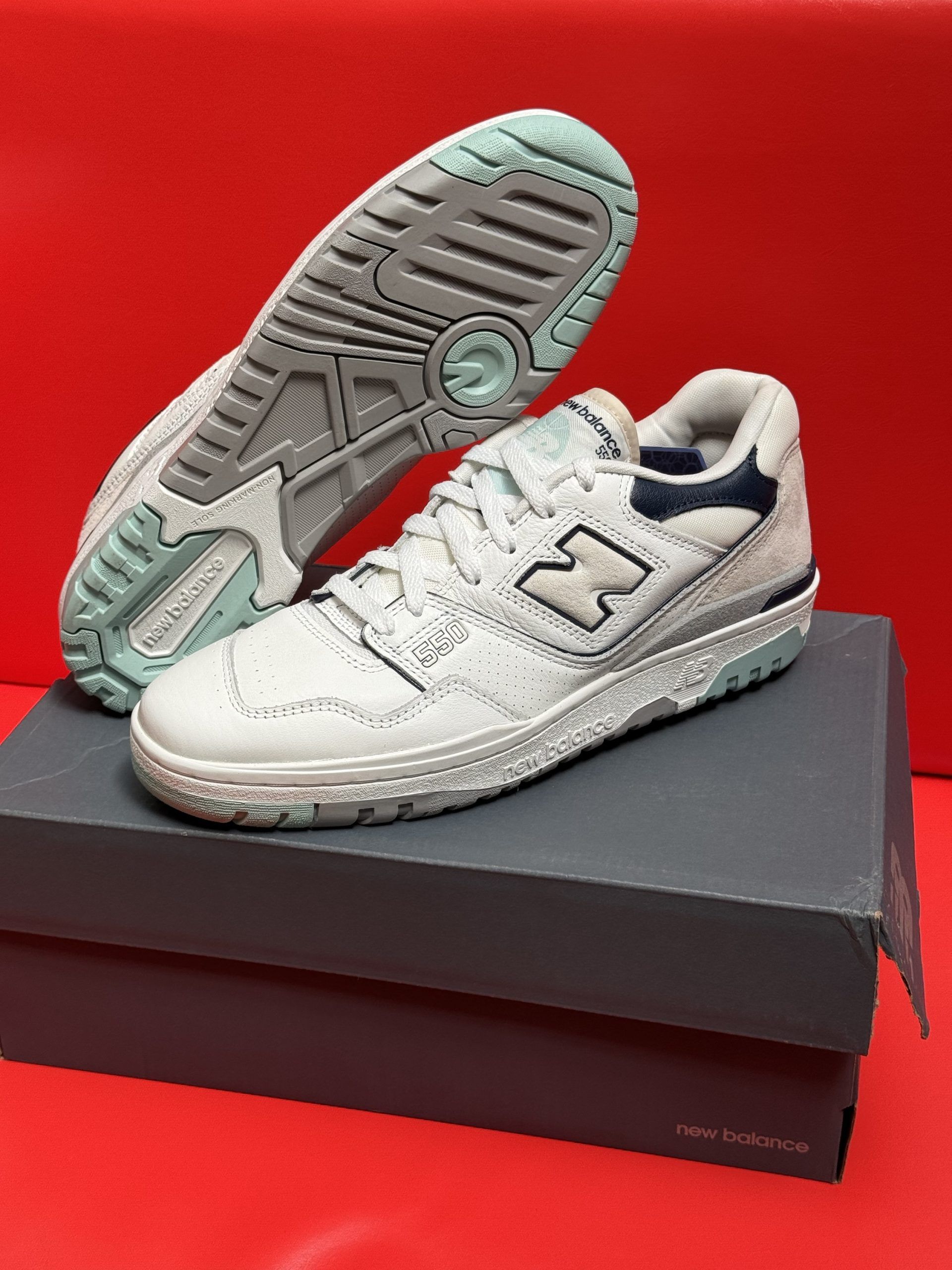 White and blue New Balance 550 sneakers on a gray box, sole visible.