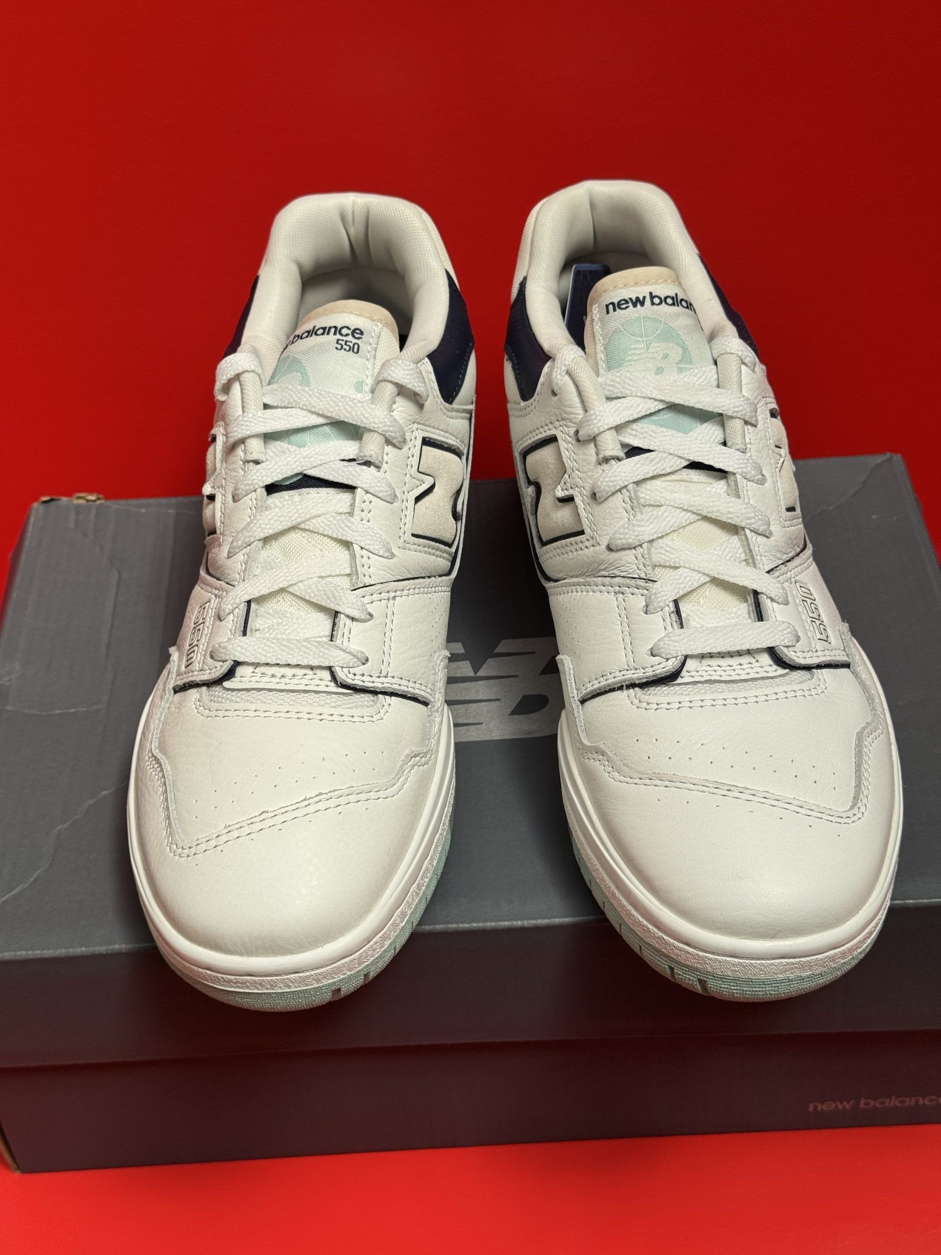 White and blue New Balance 550 sneakers on a gray box, sole visible.