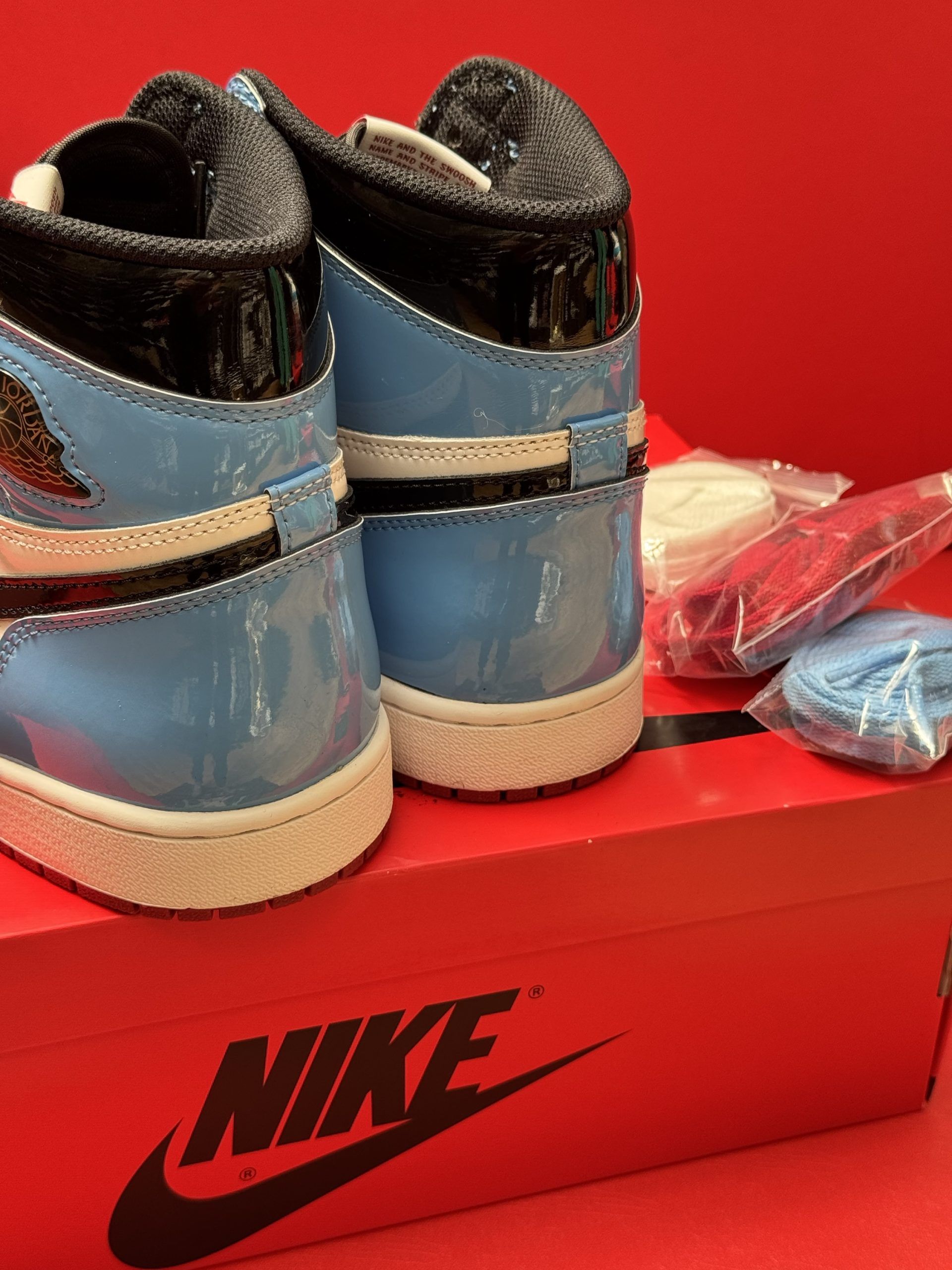 Red, white, and blue Air Jordan 1 sneaker on a red box with extra laces and a red sole against a red backdrop.