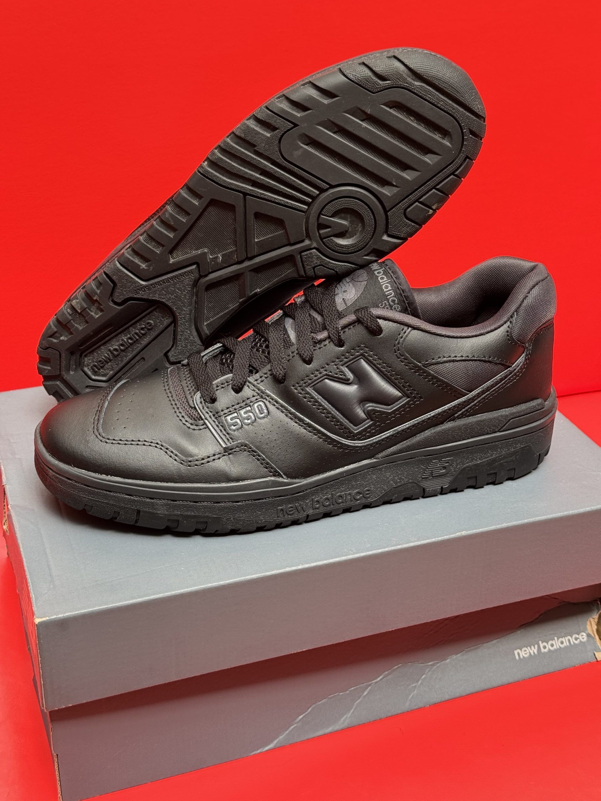 Black New Balance 550 sneakers on a box, sole up, against a red background.