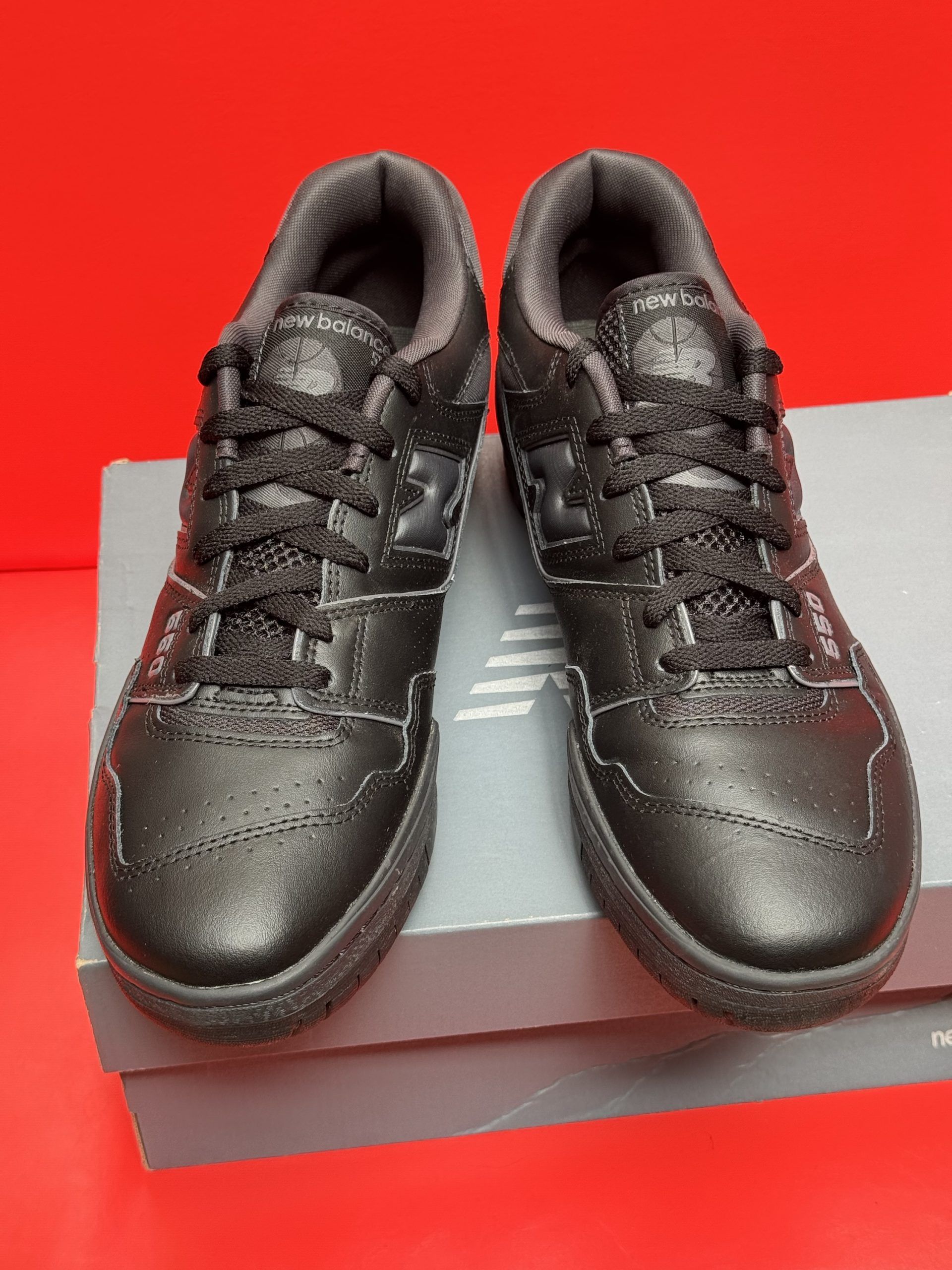 Black New Balance 550 sneakers on a box, sole up, against a red background.