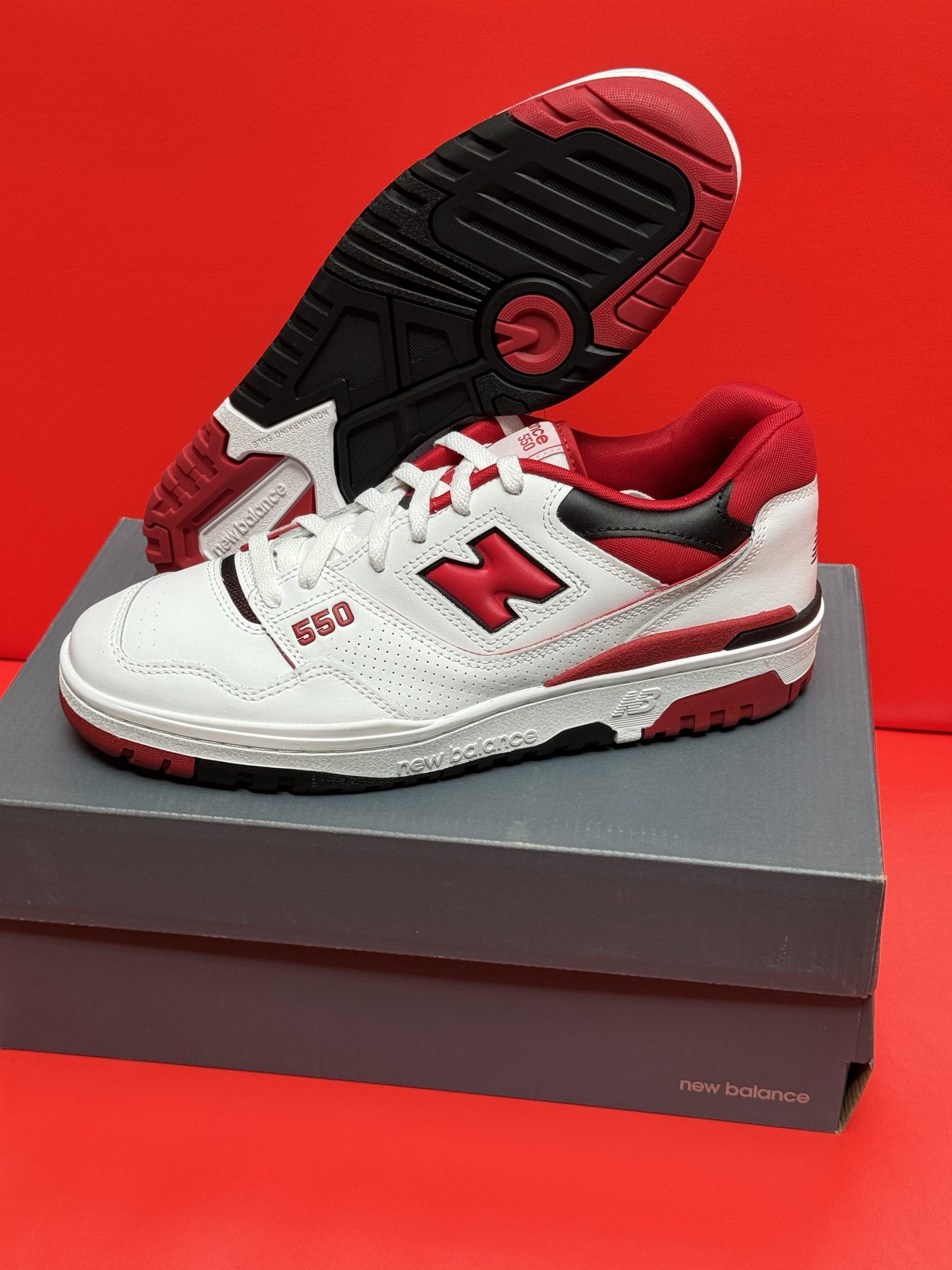 White and red New Balance 550 sneakers on a box, sole visible. Red background.