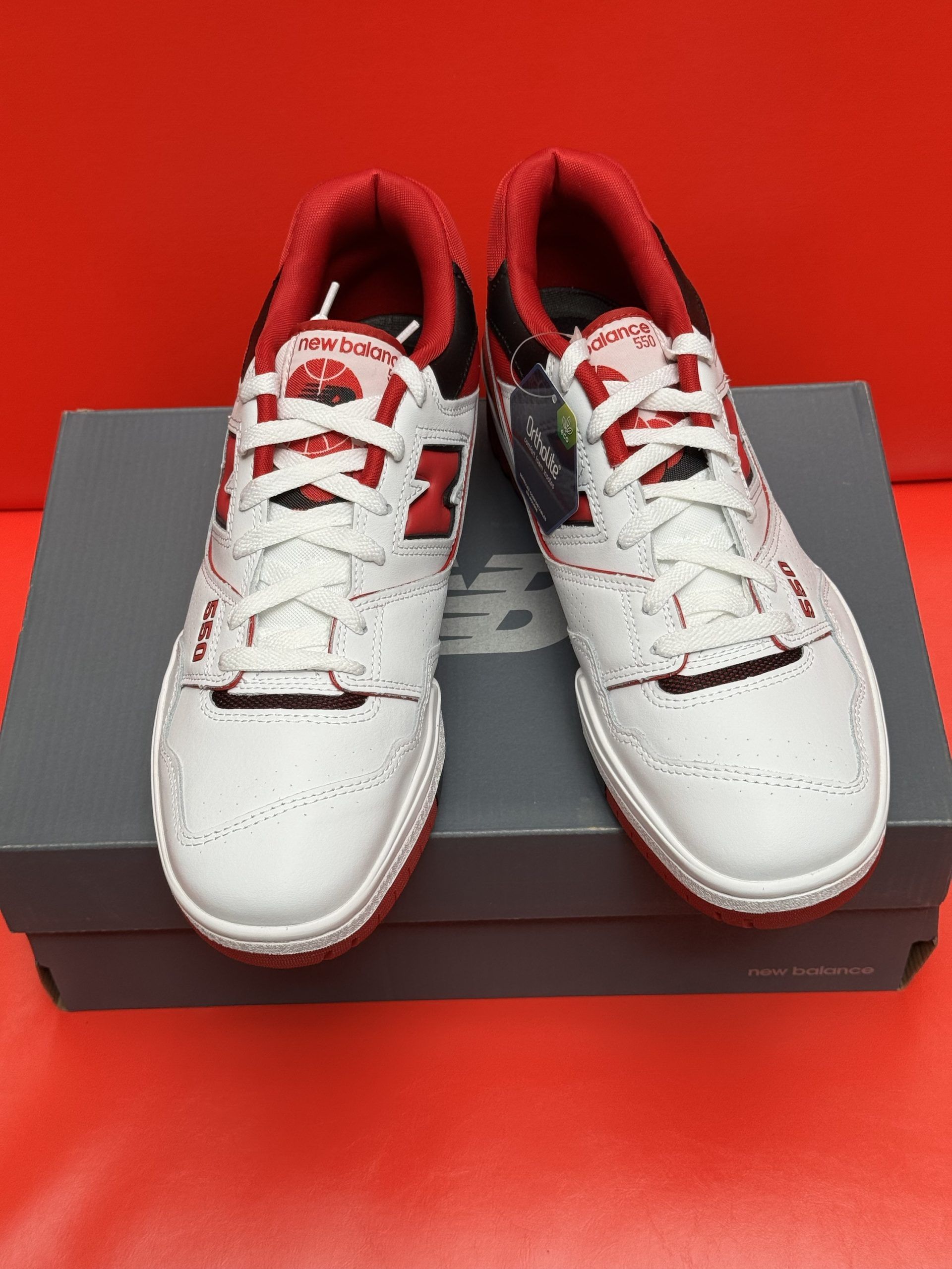White and red New Balance 550 sneakers on a box, sole visible. Red background.