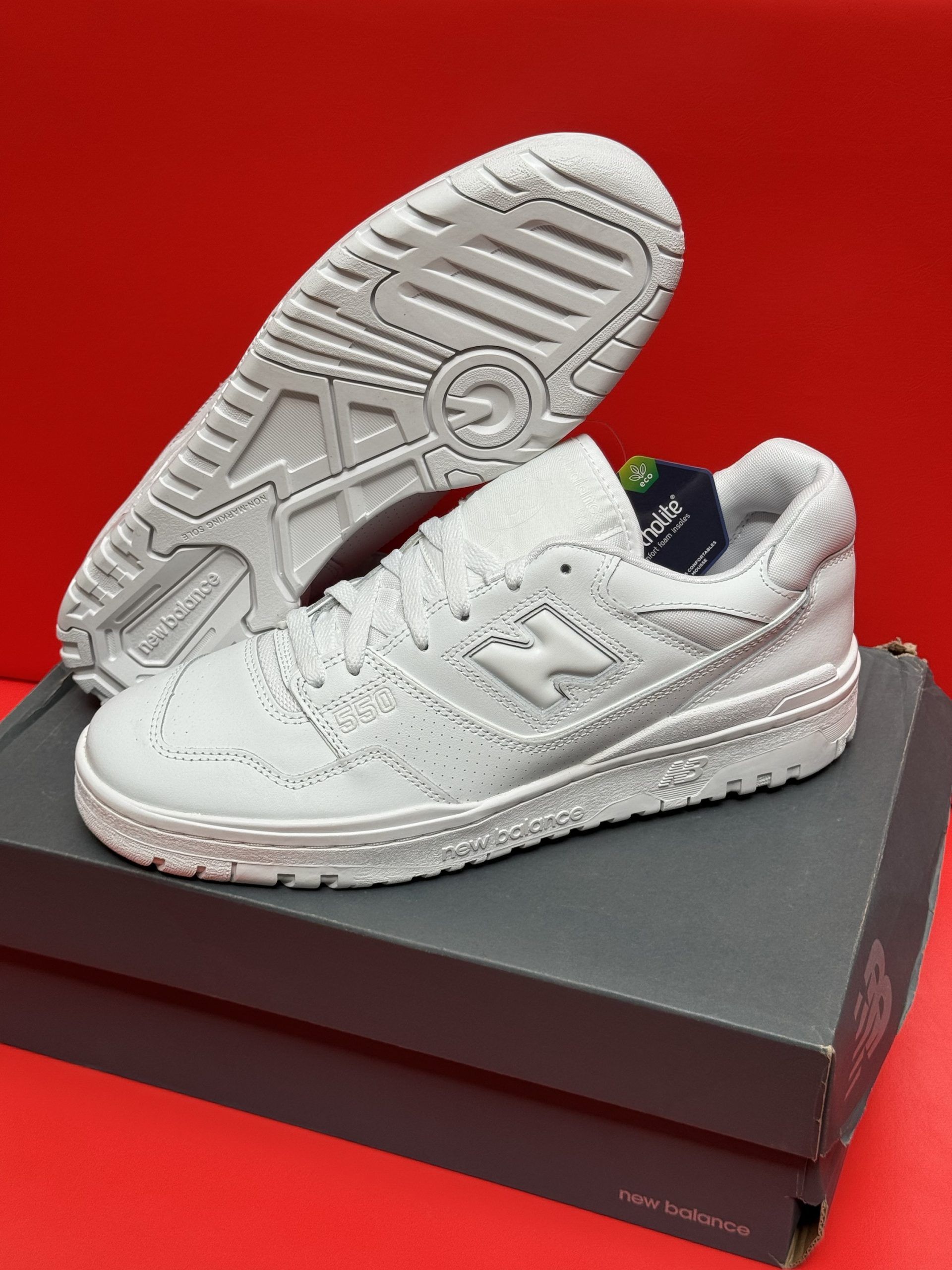 White New Balance 550 sneakers, one showing the sole, resting on a gray box against a red background.