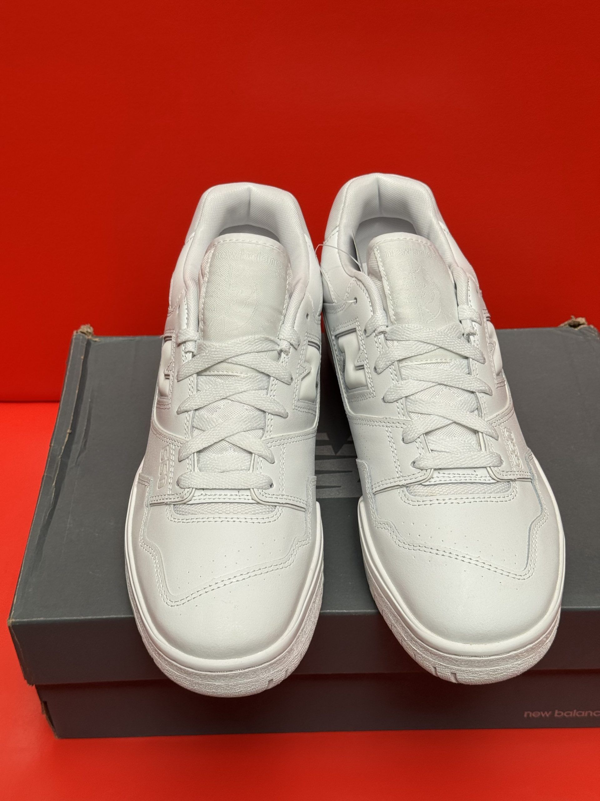 White New Balance 550 sneakers, one showing the sole, resting on a gray box against a red background.