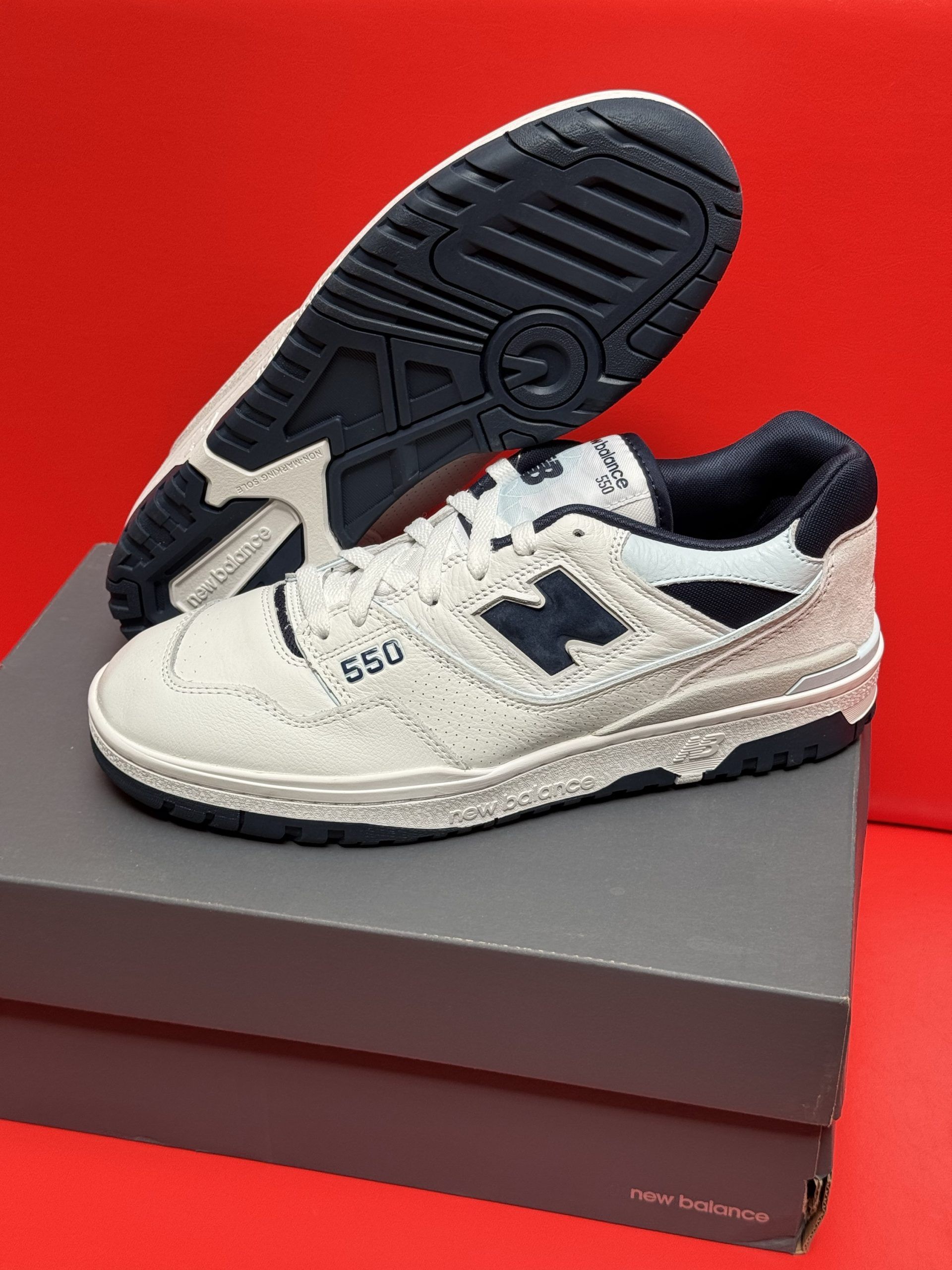 New Balance 550 sneakers with off-white and navy accents, sitting on a gray shoebox. Sole is visible.
