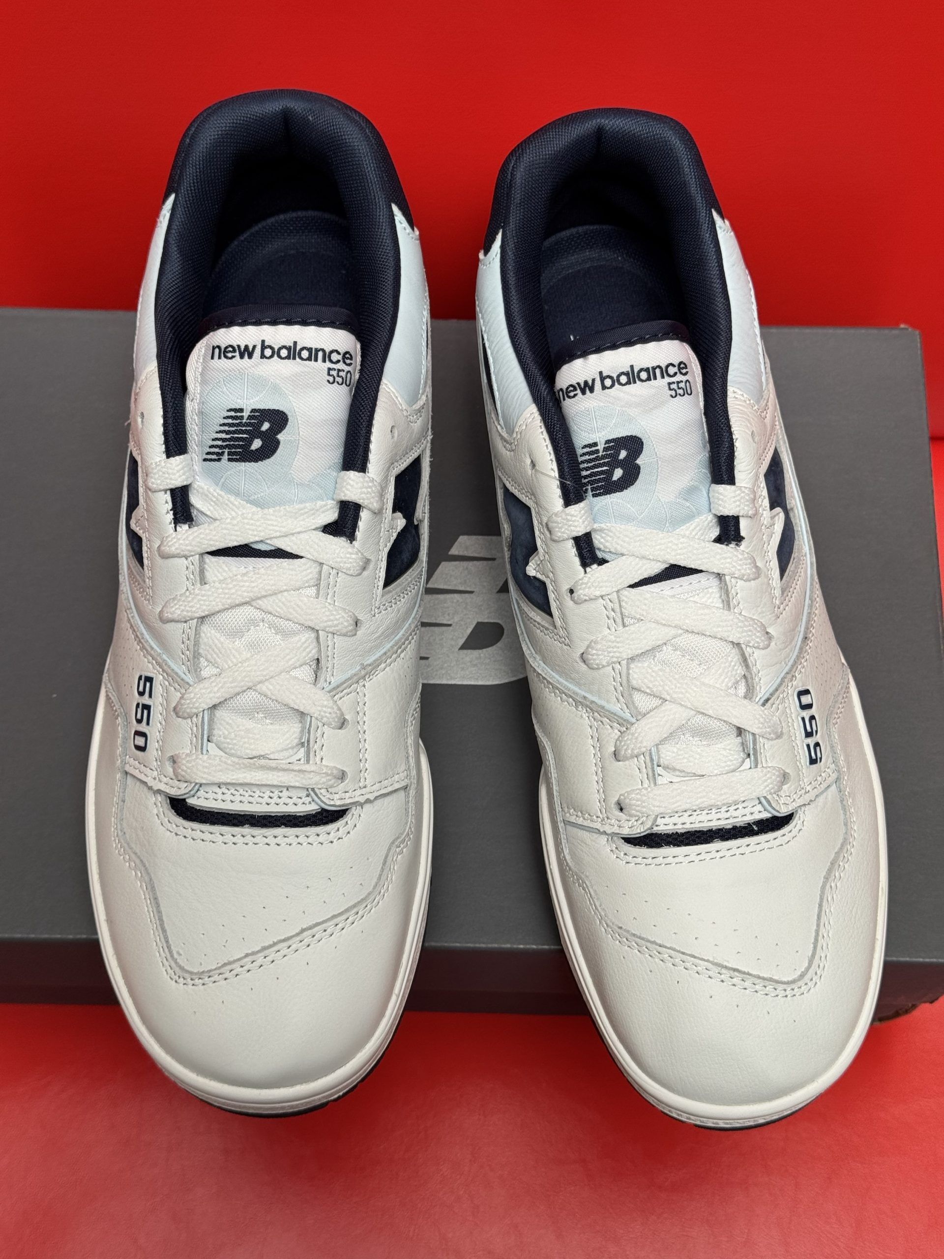 New Balance 550 sneakers with off-white and navy accents, sitting on a gray shoebox. Sole is visible.