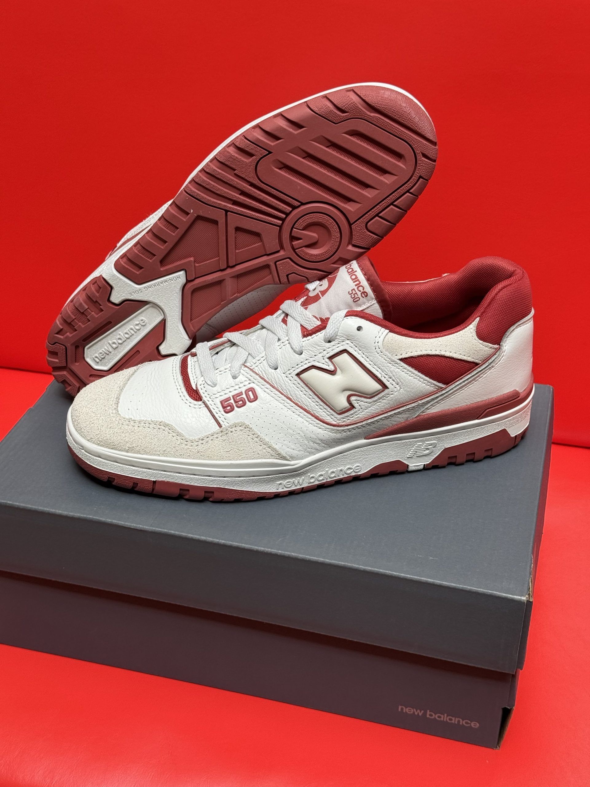 White and red New Balance 550 sneakers on a gray shoebox, against a red background. One shoe is upside down, showing the sole.
