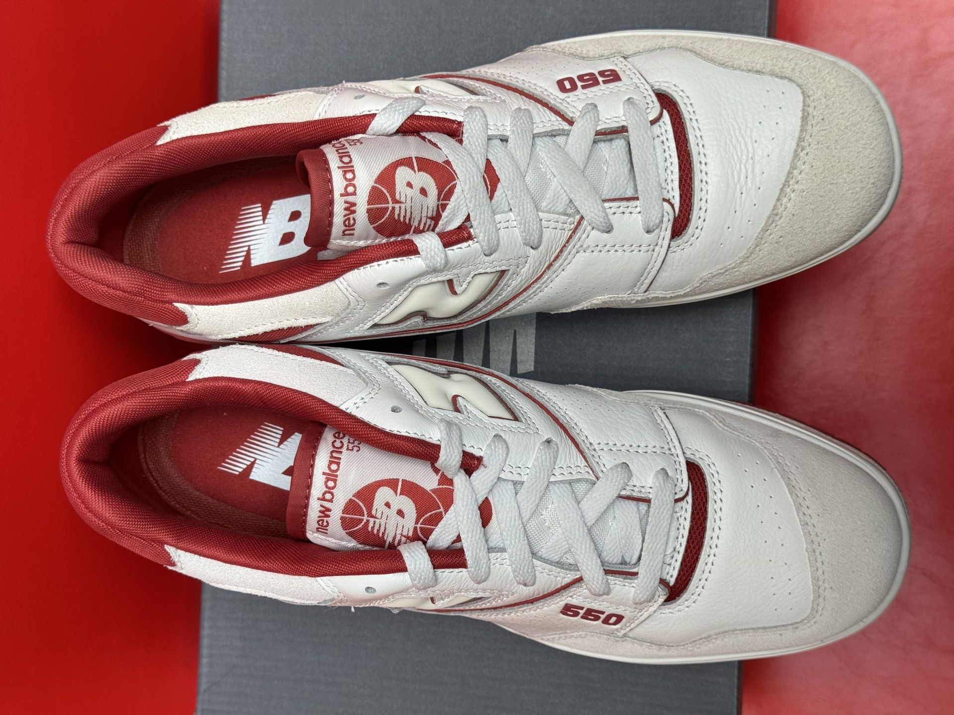 White and red New Balance 550 sneakers on a gray shoebox, against a red background. One shoe is upside down, showing the sole.