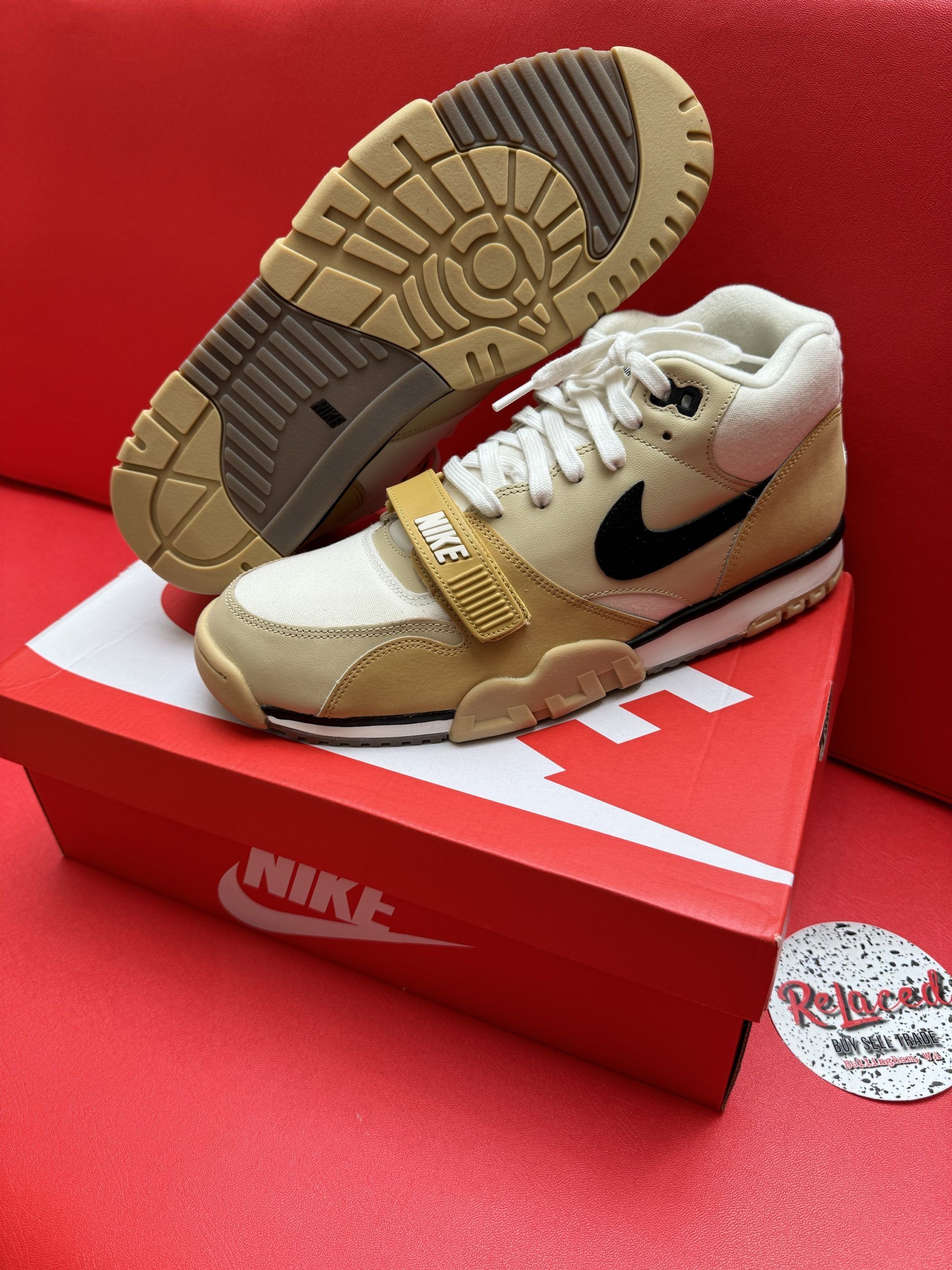Beige and black Nike Air Trainer sneakers on a red Nike box with a beige sole, against a red surface.