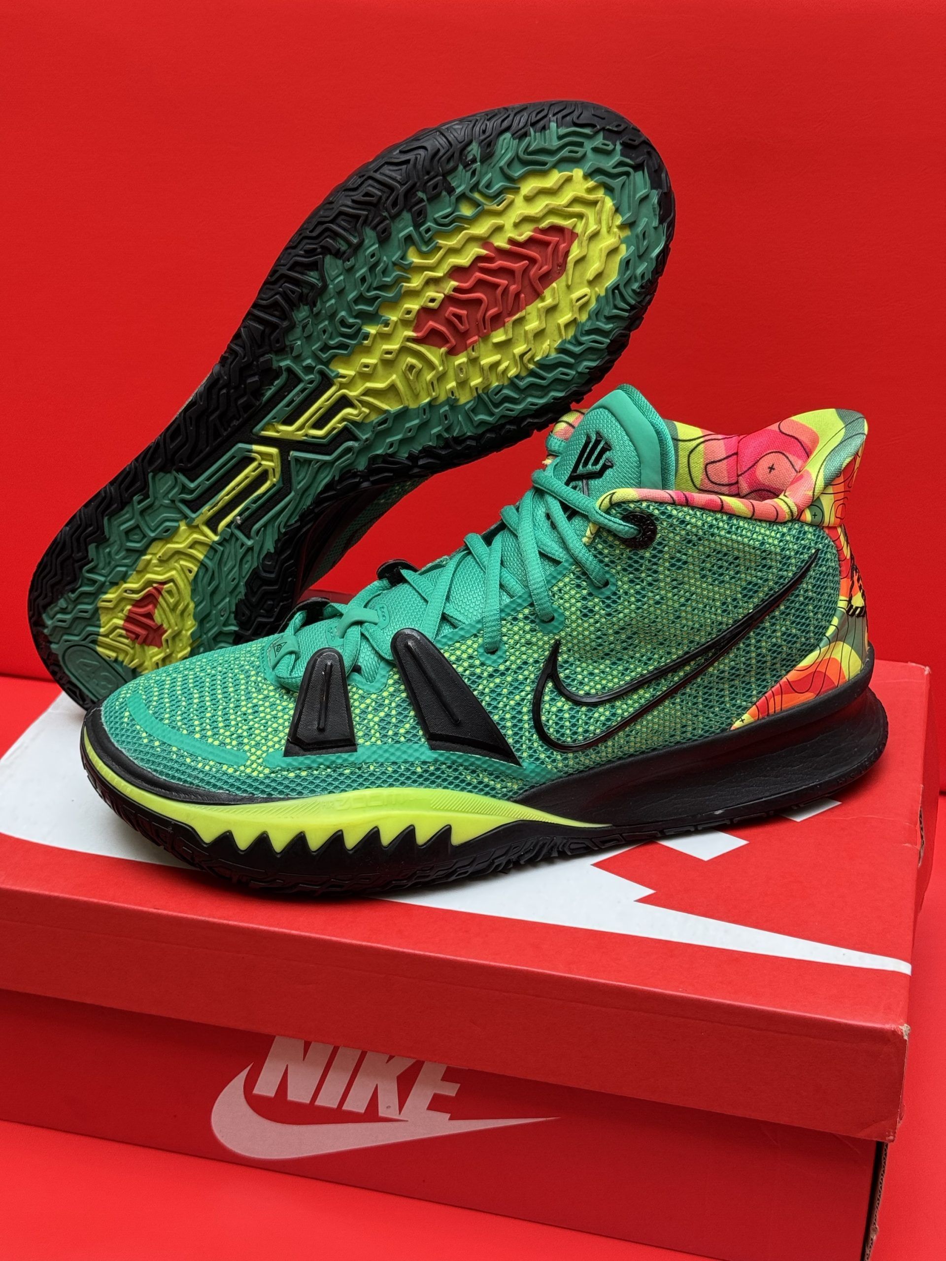 Green and black Nike Kyrie 7 basketball shoe on a box, sole visible, red background.