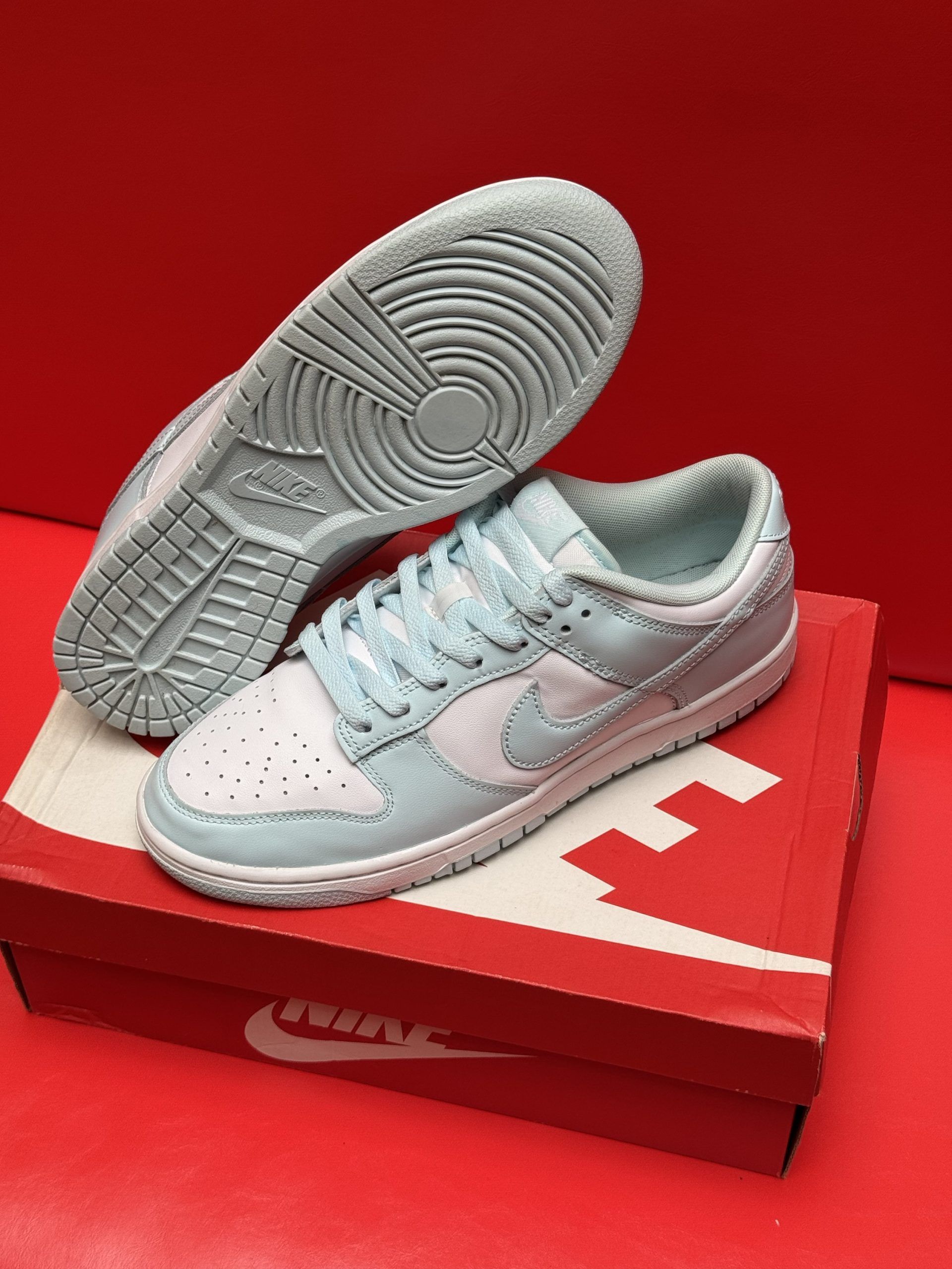Light blue Nike Dunk Low sneakers on a red Nike box, against a red background. One sneaker’s sole is visible.