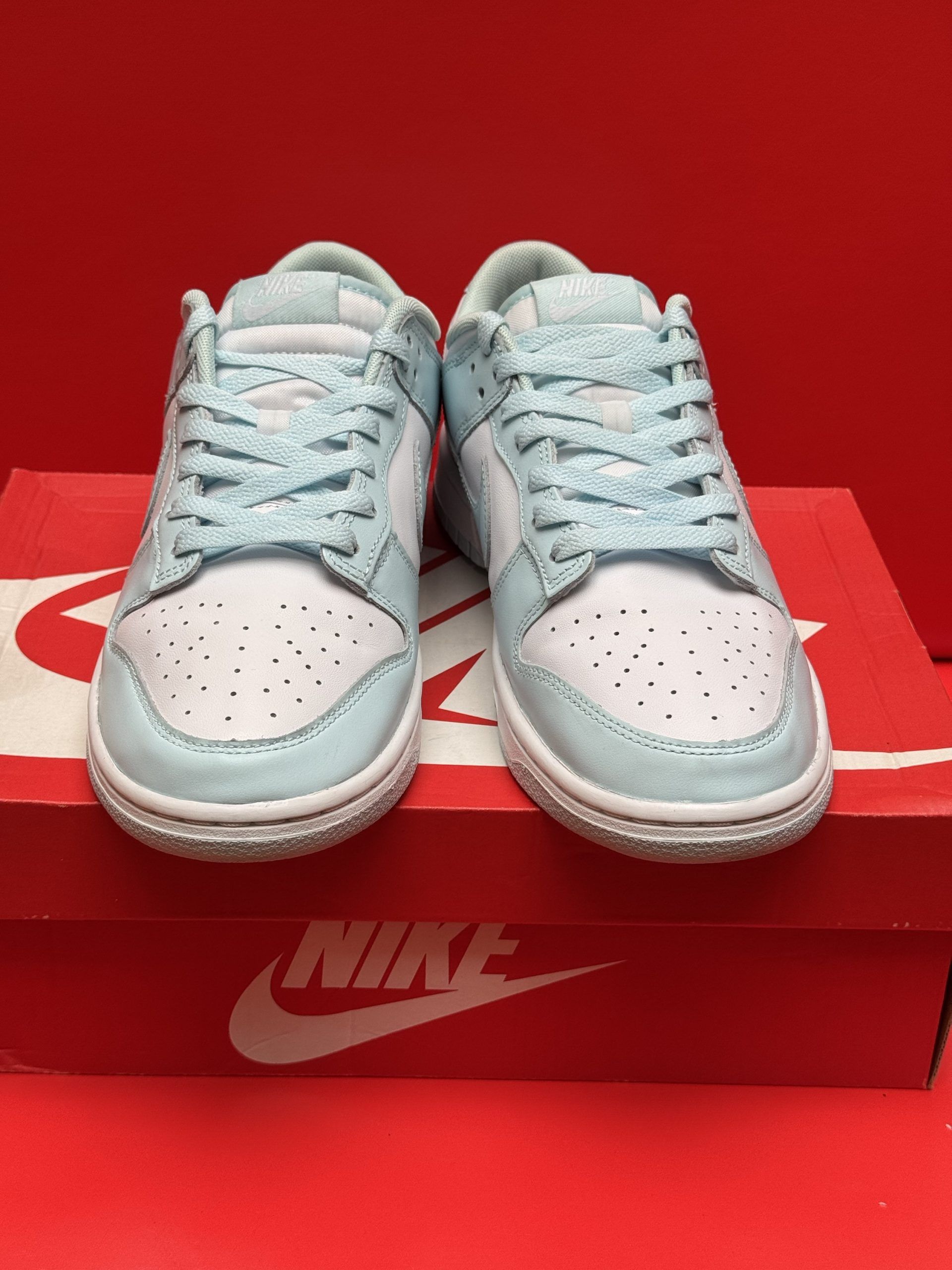 Light blue Nike Dunk Low sneakers on a red Nike box, against a red background. One sneaker’s sole is visible.