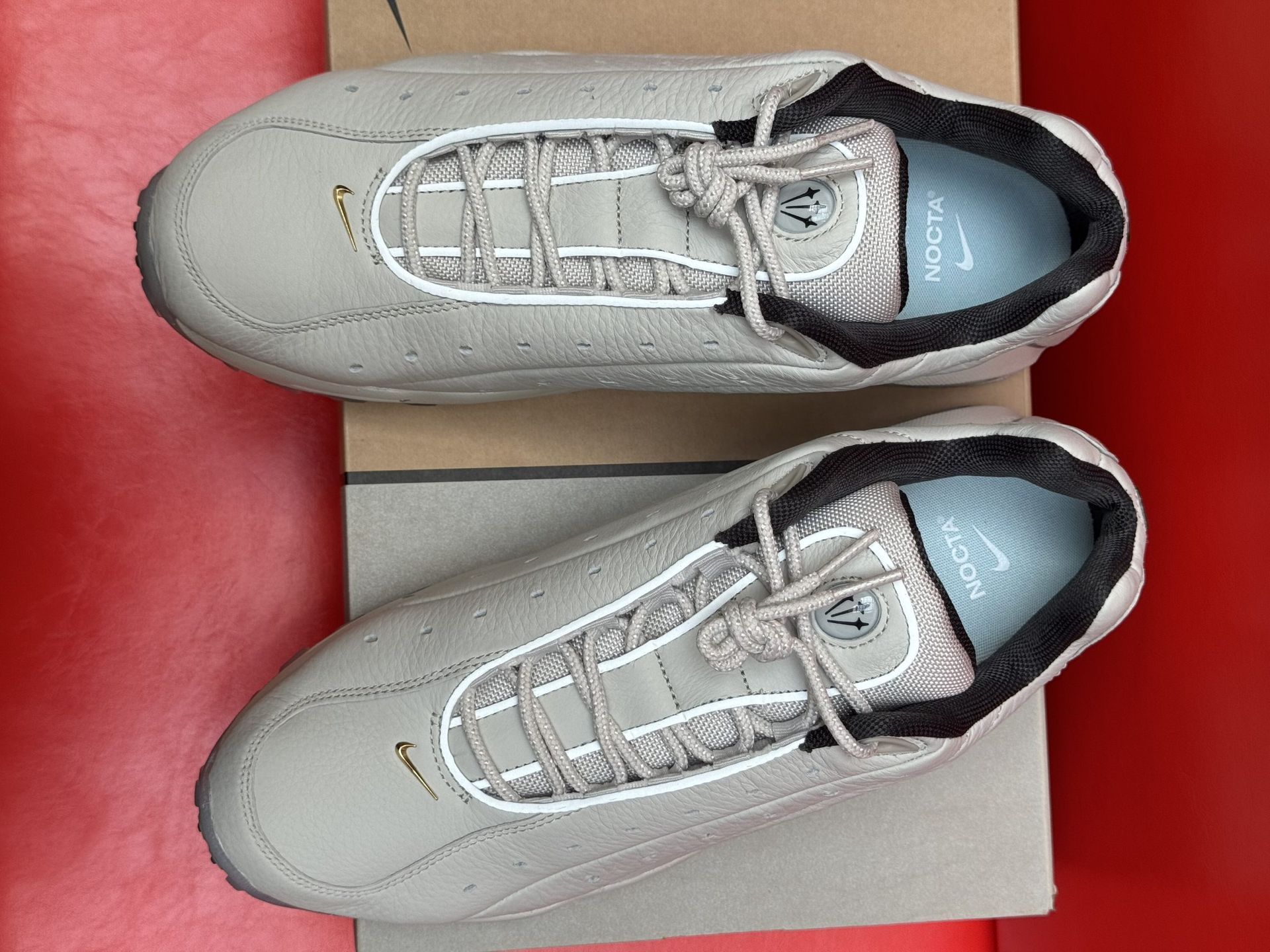 Beige Nike Air Max sneakers on box with sole visible, set against a red background.