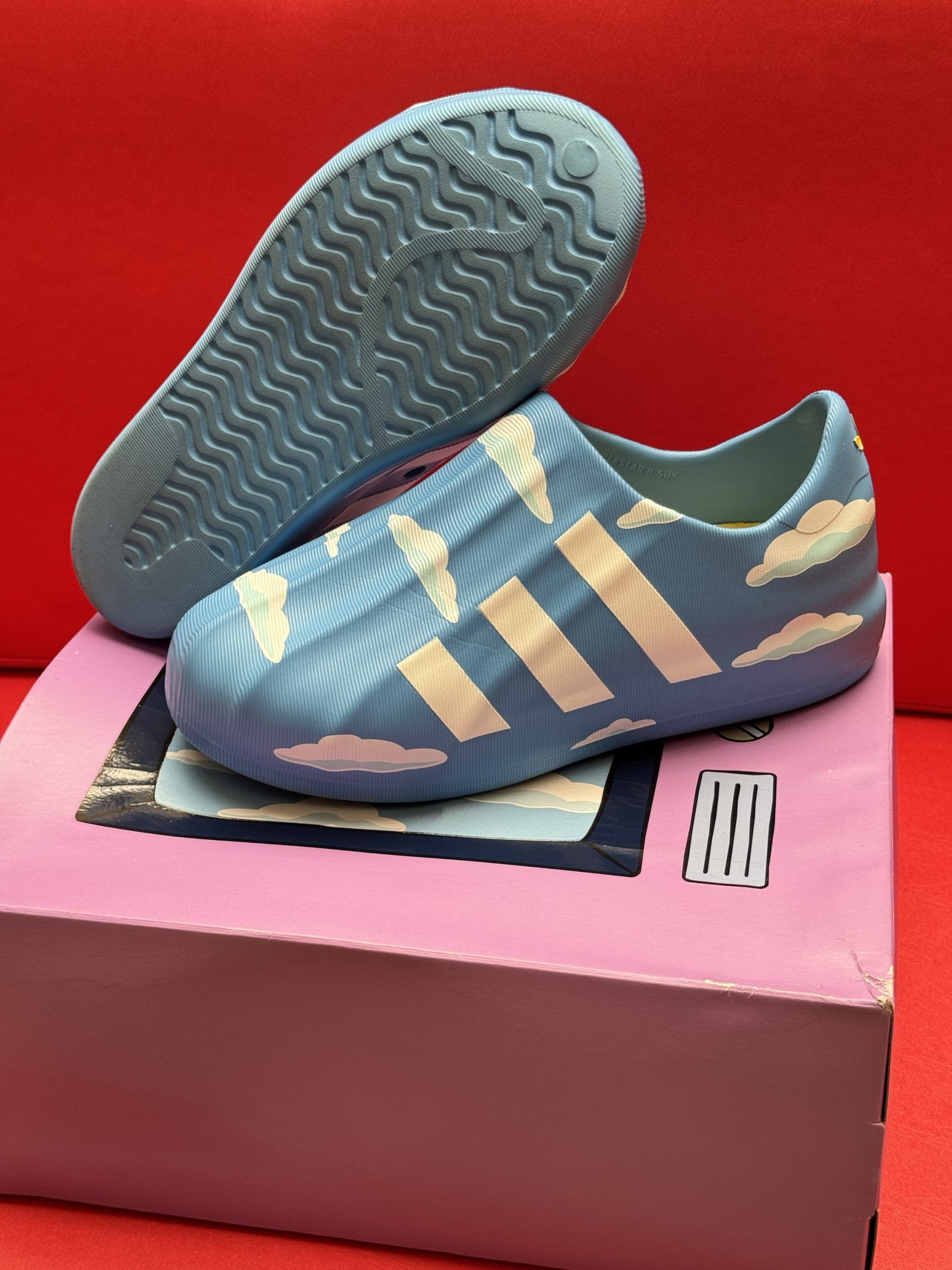 Blue and white cloud-patterned slip-on shoes on a pink box, set against a red background.