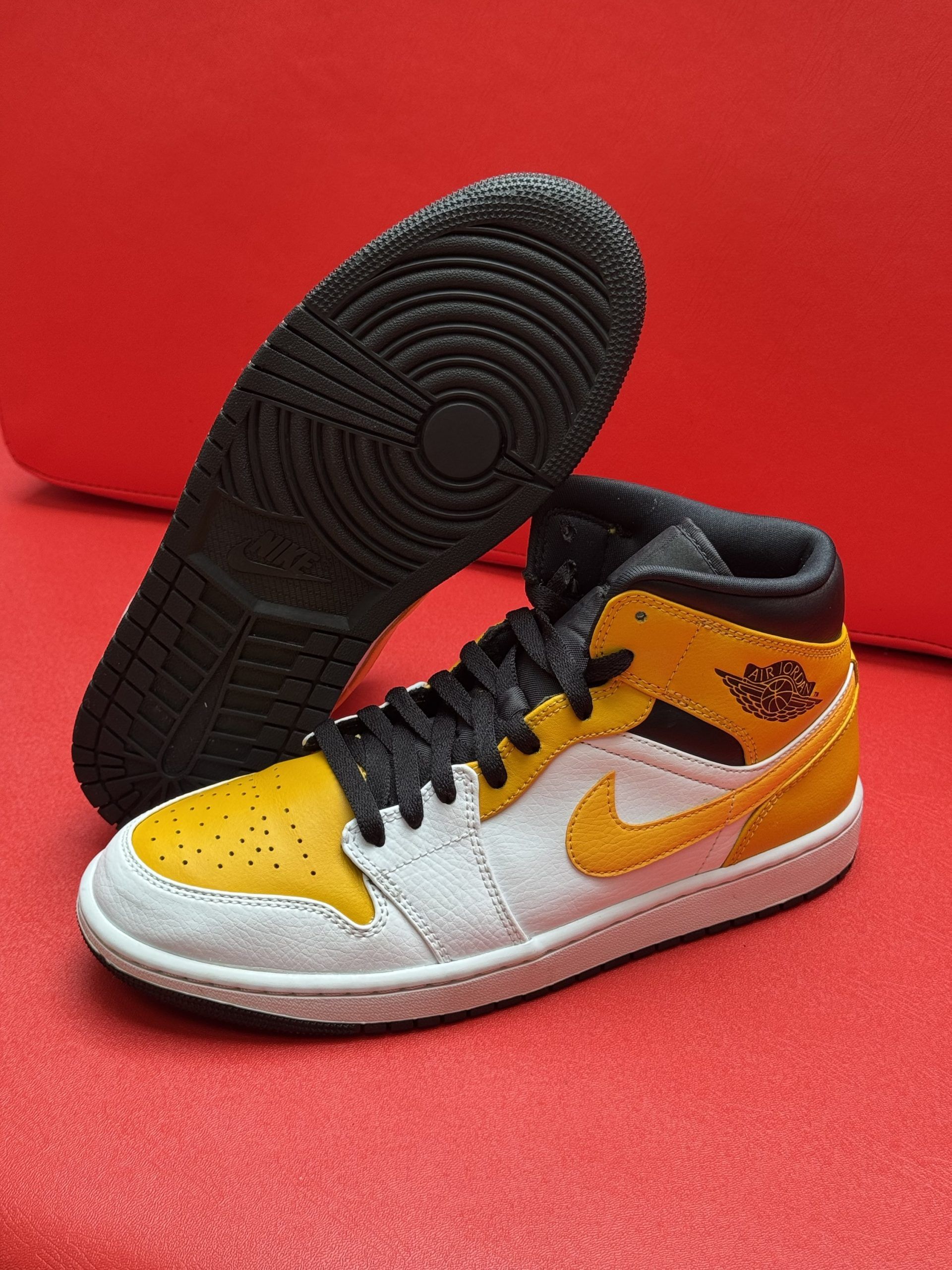 Yellow, white, and black Air Jordan 1 sneaker with black sole on red surface.