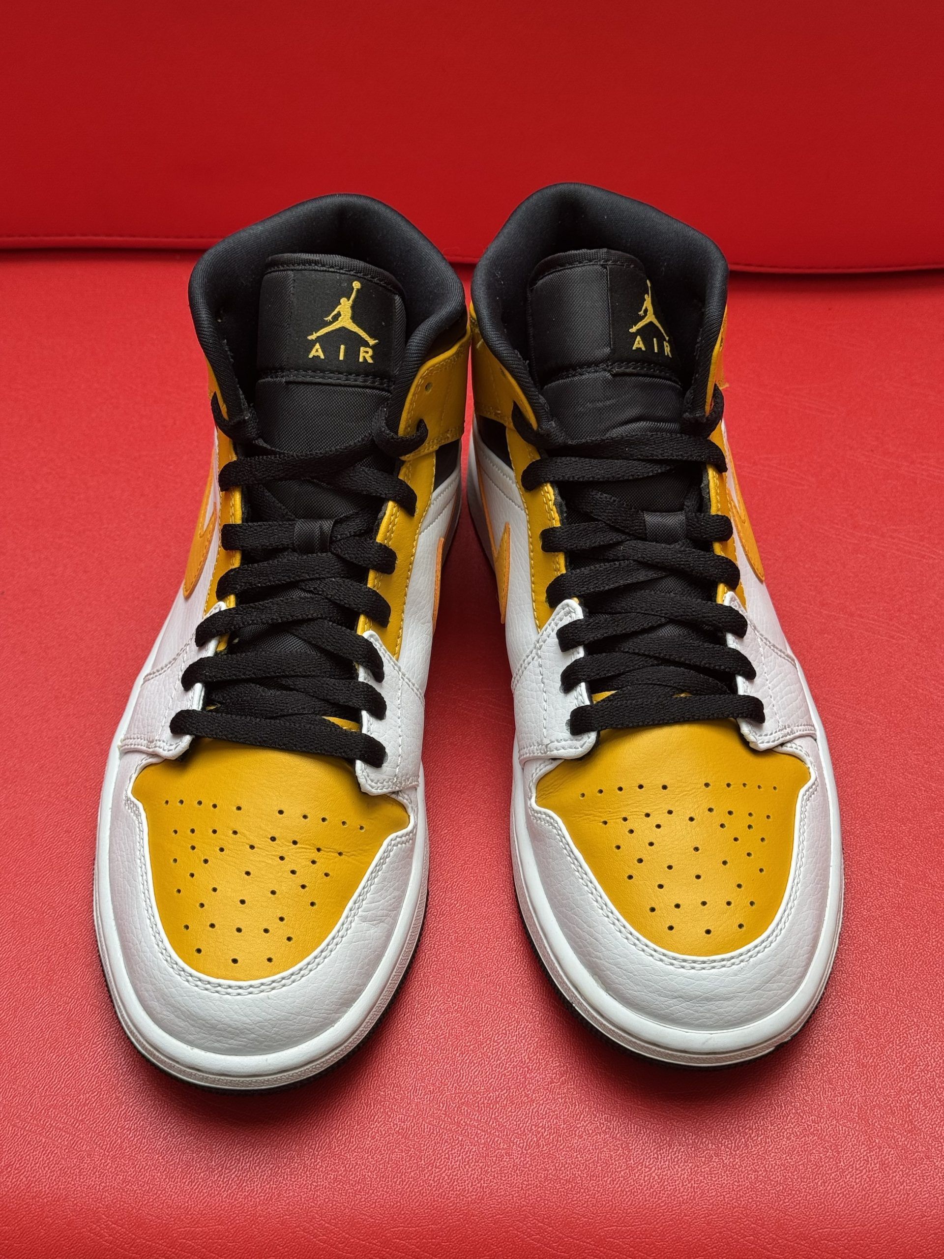 Yellow, white, and black Air Jordan 1 sneaker with black sole on red surface.