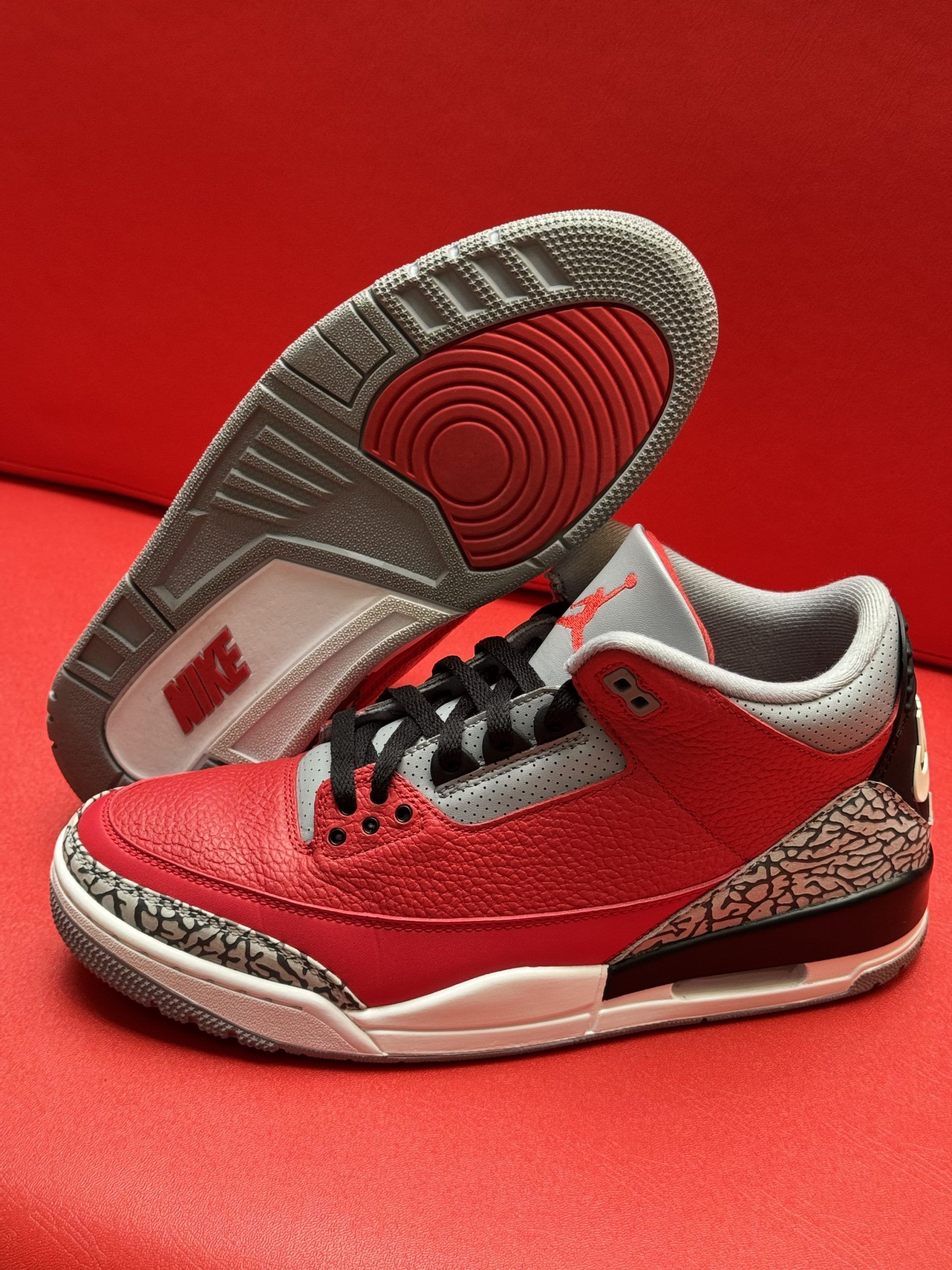 Red and gray Air Jordan 3 sneaker on a red surface, with a visible sole.