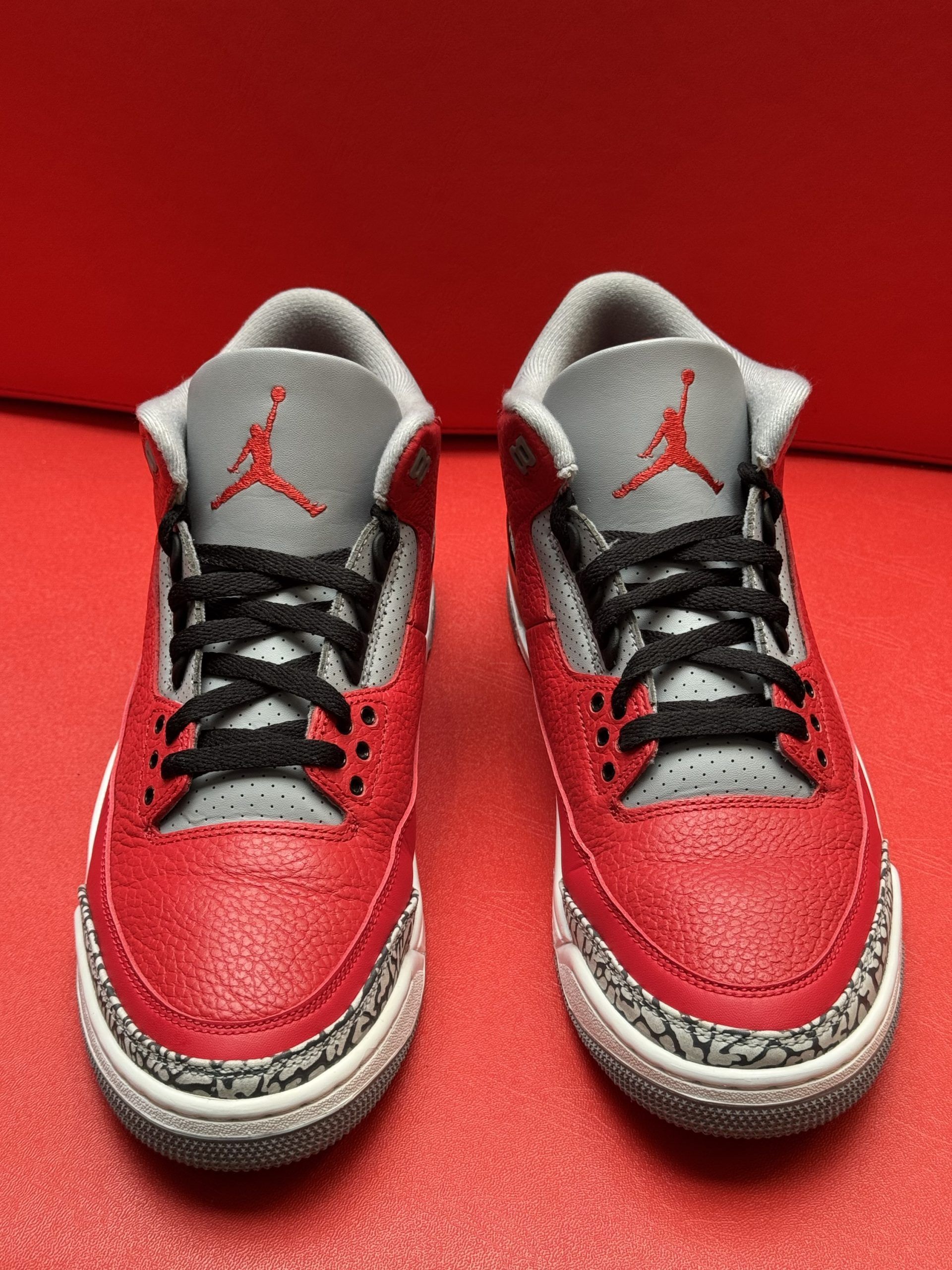 Red and gray Air Jordan 3 sneaker on a red surface, with a visible sole.