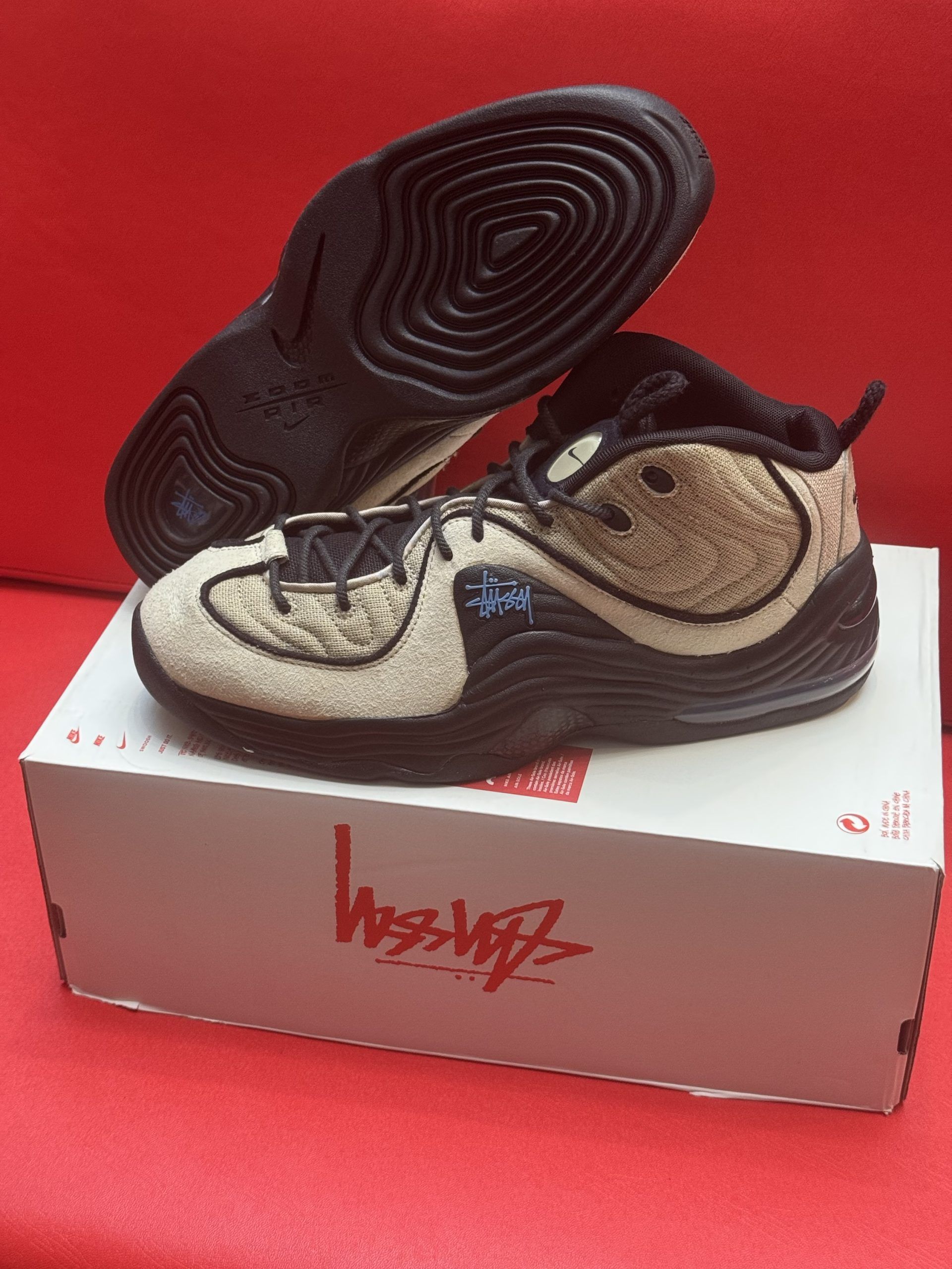Tan and black Nike Penny Hardaway sneakers on a box with the Stussy logo, red background.