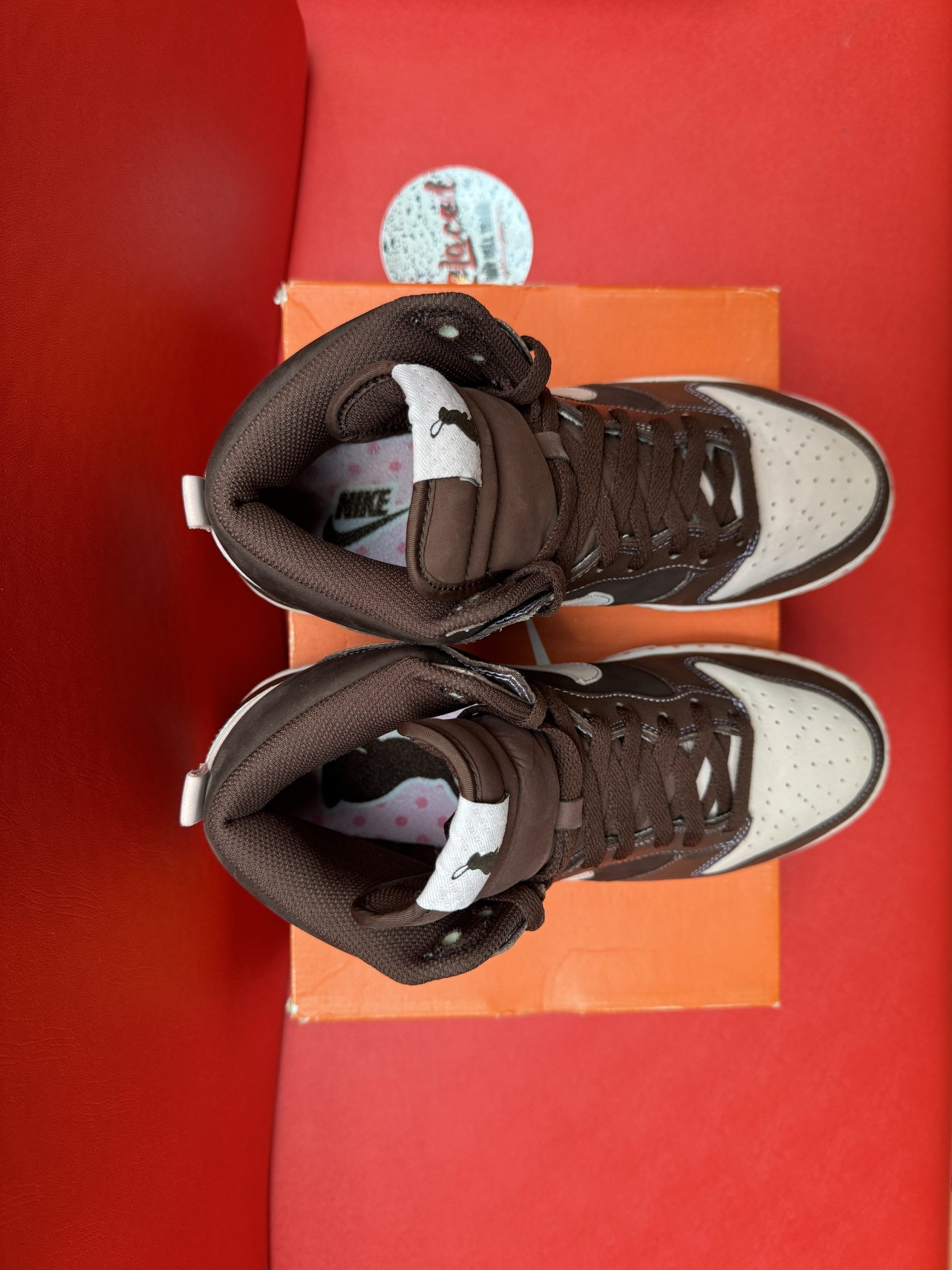 Nike Dunk High sneakers, brown and cream, on an orange box, brown sole, white swoosh on a red surface.
